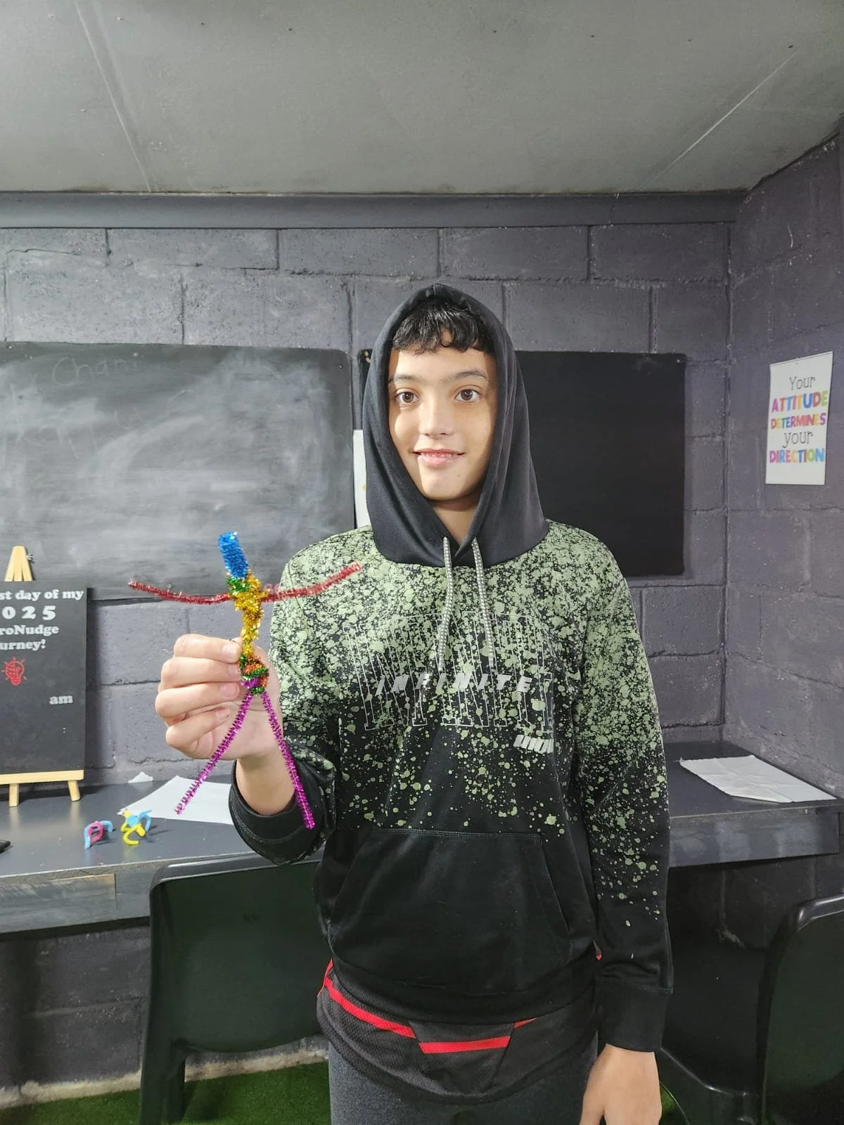 Young boy in a black and green hoodie holding a colorful pipe cleaner craft figure, standing in a room with a chalkboard and motivational posters in the background.