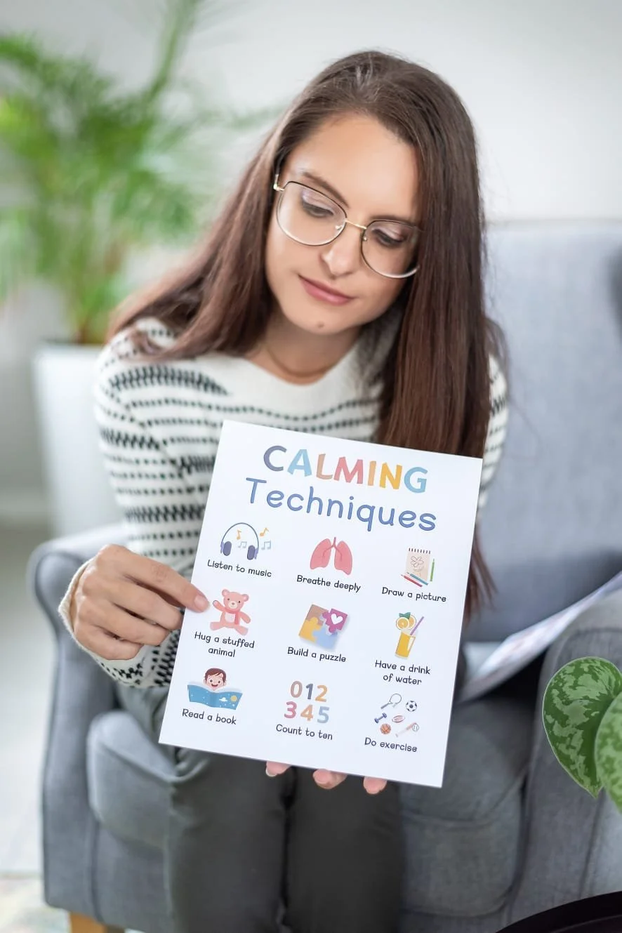 A young woman with glasses and long brown hair sitting on a gray couch, holding a sign titled 'Calming Techniques' with illustrations and tips for relaxation.