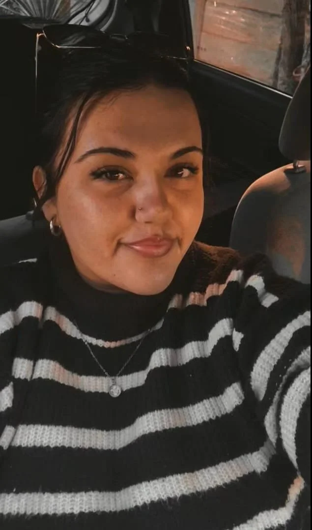 A woman with dark hair and freckles sitting in a car, wearing a black and white striped sweater, sunglasses on her head, earrings, and a necklace, smiling at the camera.