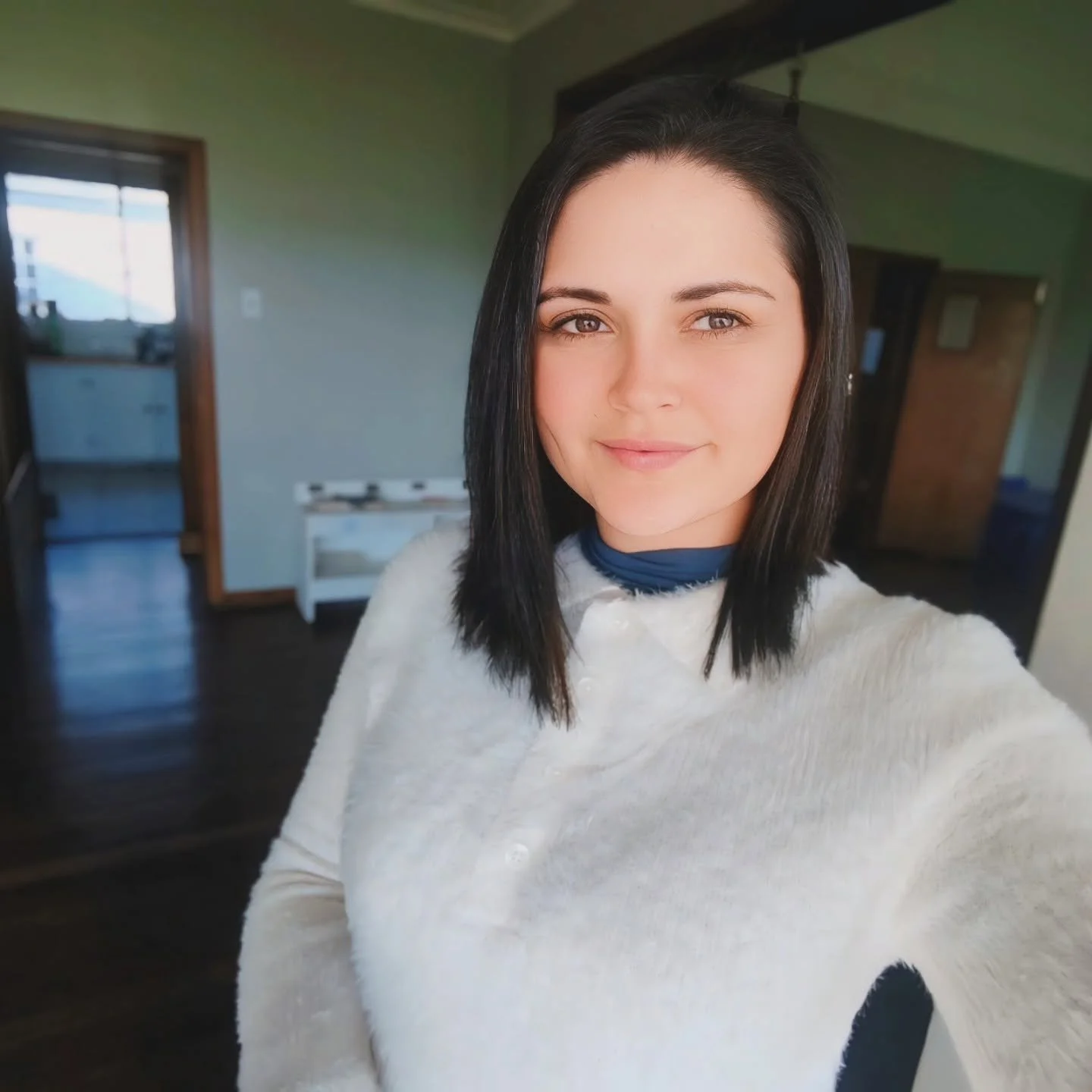 A woman with shoulder-length black hair and fair skin taking a selfie in a room with green walls and dark hardwood floors, with a window and door in the background.