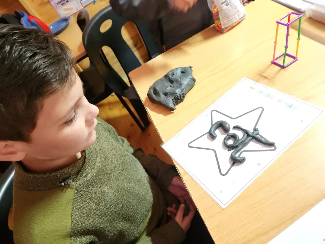 A young boy is sitting at a wooden table looking at a sheet of paper with a star outline and the word 'pat' written with Play-Doh on it. There is a rock and a colorful geometric figure on the table.