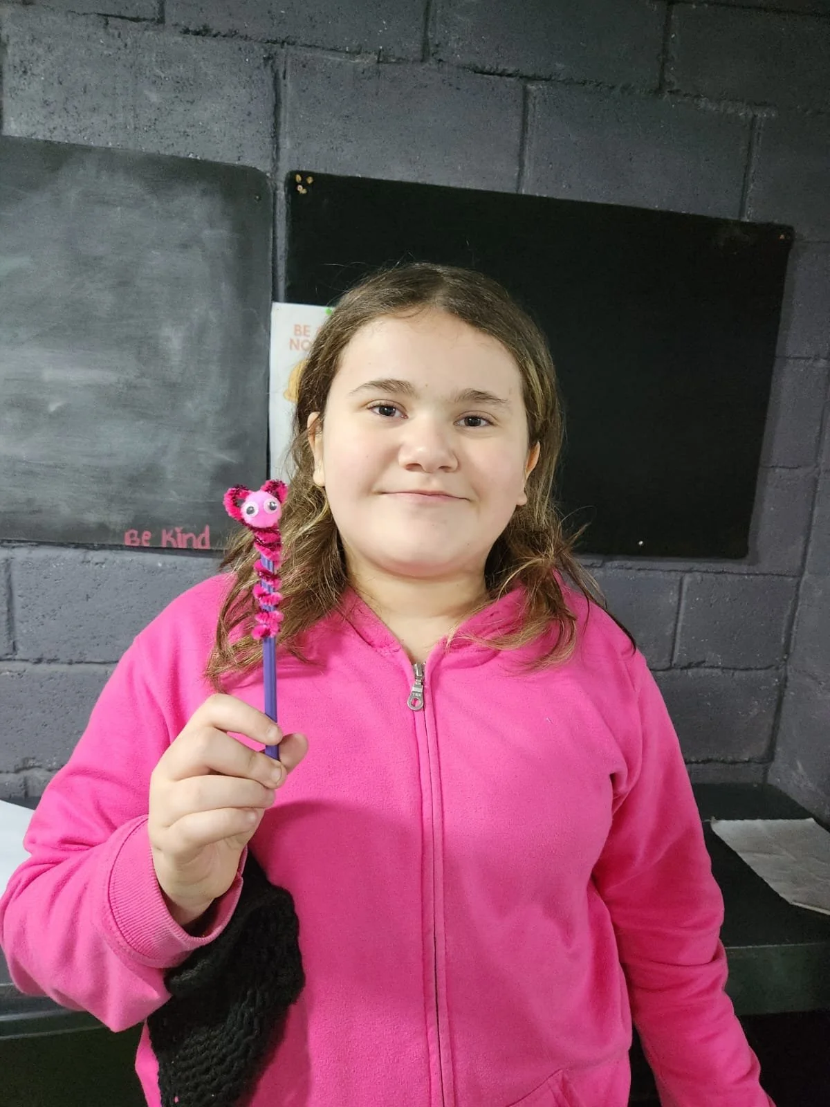 A young girl wearing a pink jacket holding a pink fuzzy pen with a monster face. She is standing in front of a gray brick wall with chalkboards, one of which has the words 'Be Kind' written on it.