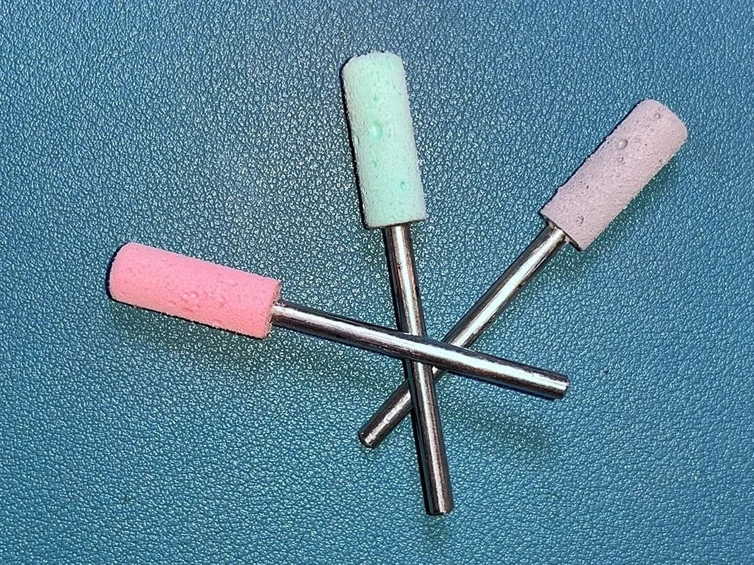 Drei Schleifstifte mit bunten Kunststoffhülsen in den Farben pink, grün und grau, auf einer blauen Oberfläche angeordnet.