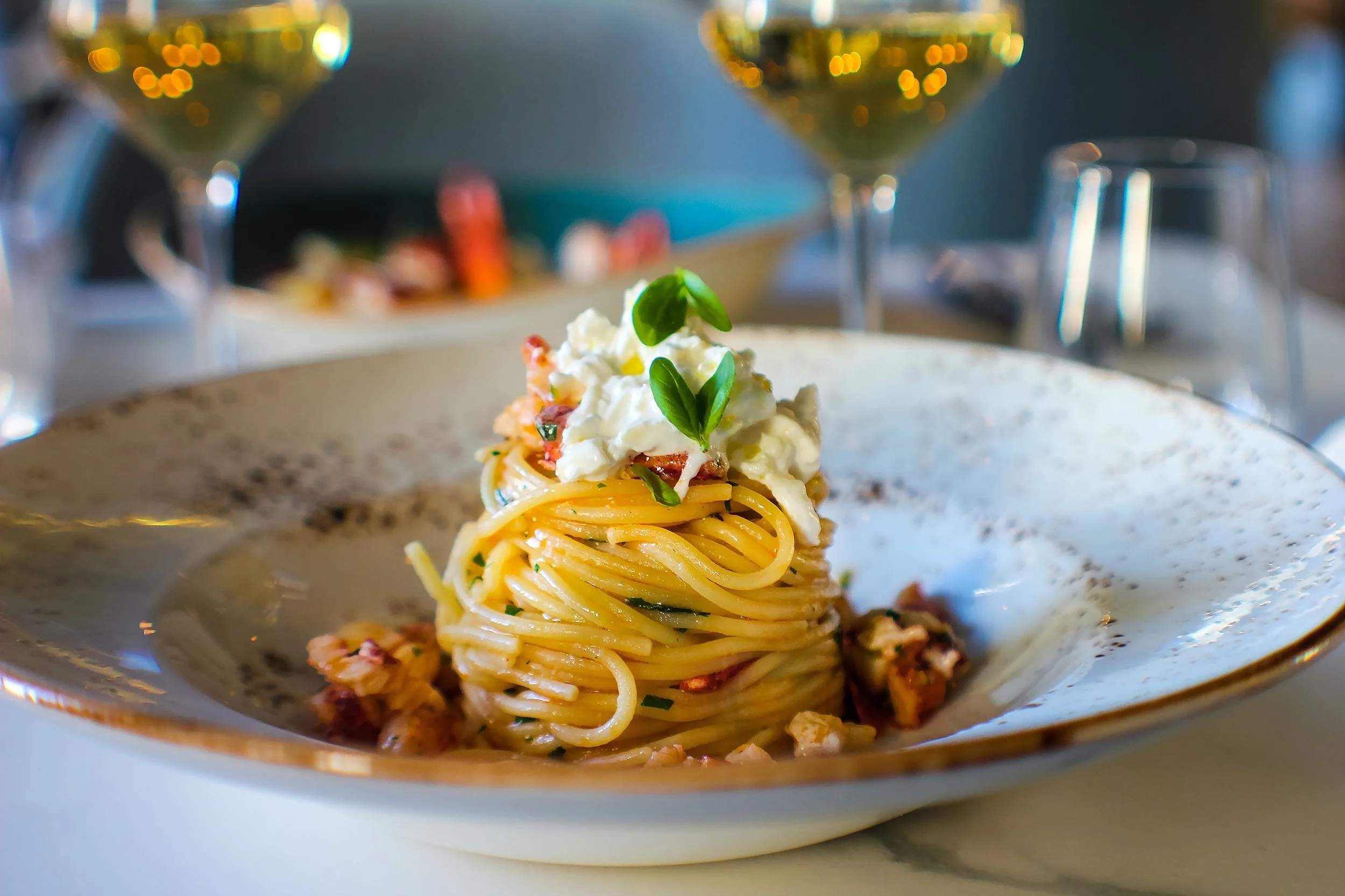 Close-up of a plate of spaghetti pasta topped with ricotta cheese and fresh basil leaves, with blurred wine glasses in the background.