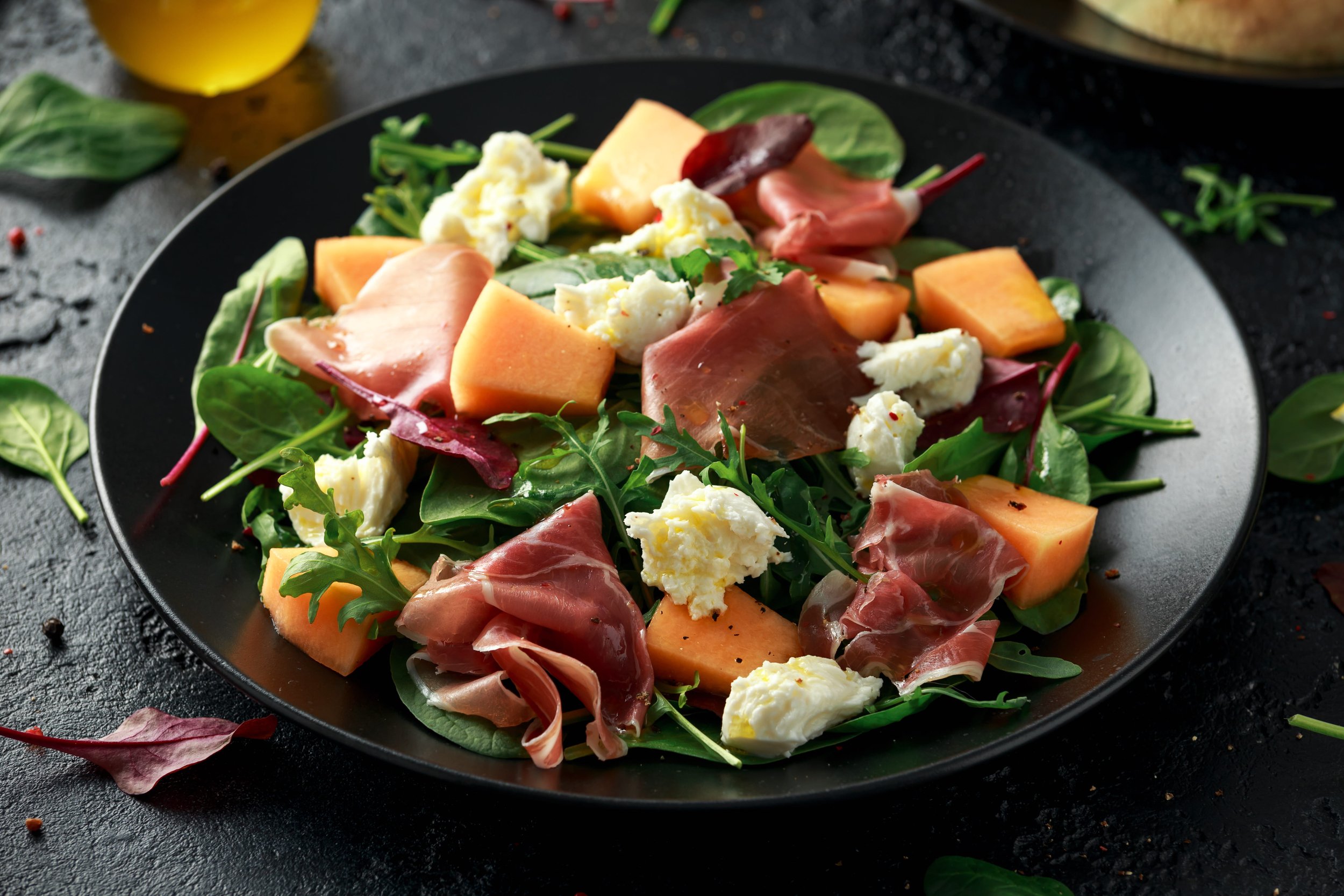 A fresh salad with mixed greens, slices of cantaloupe, crumbled cheese, and slices of prosciutto on a black plate, on a dark textured surface with scattered leaves and a glass of yellow beverage in the background.