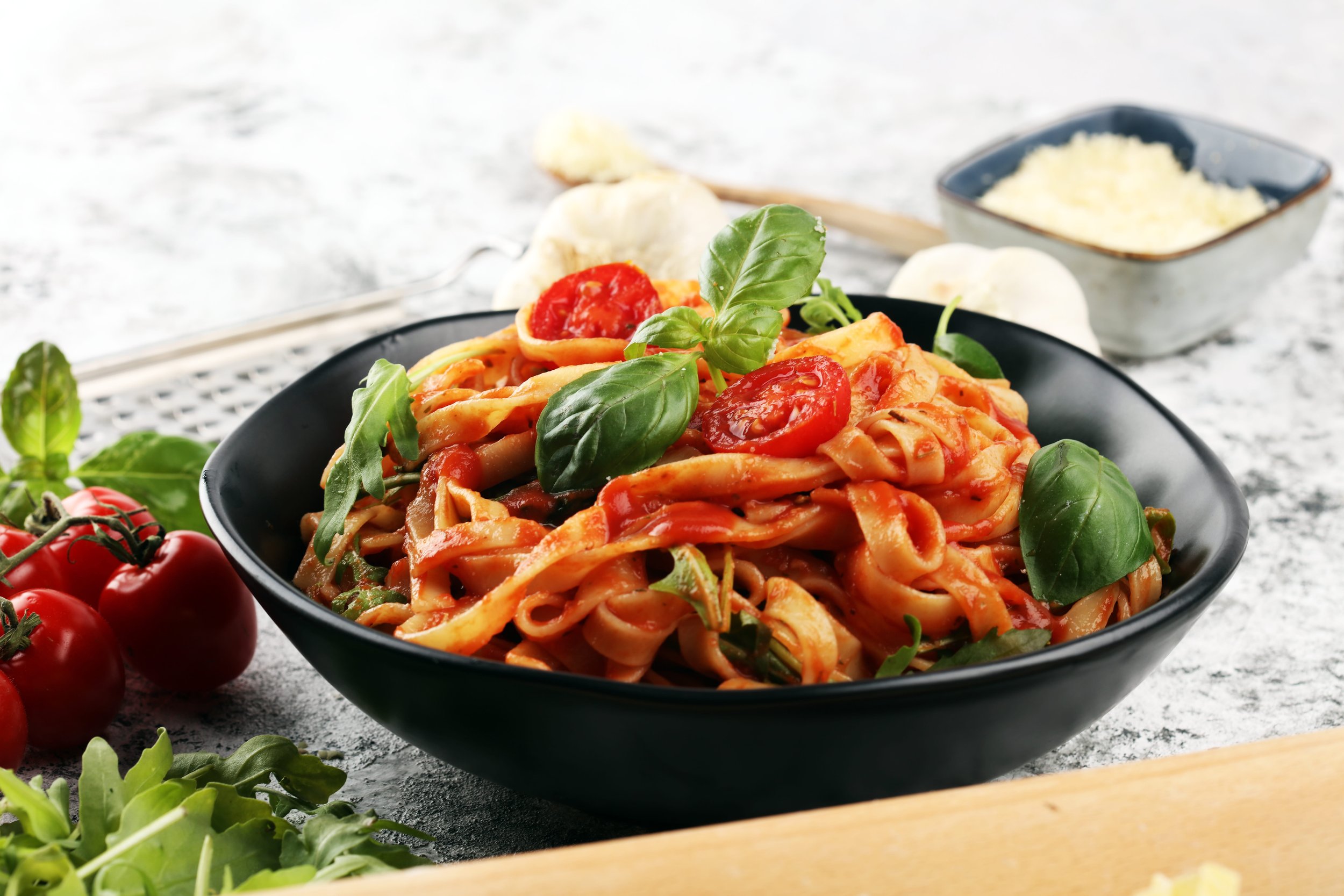 A bowl of fettuccine pasta with tomato sauce, garnished with fresh basil leaves. Surrounding it are cherry tomatoes, garlic, and green herbs on a gray surface.