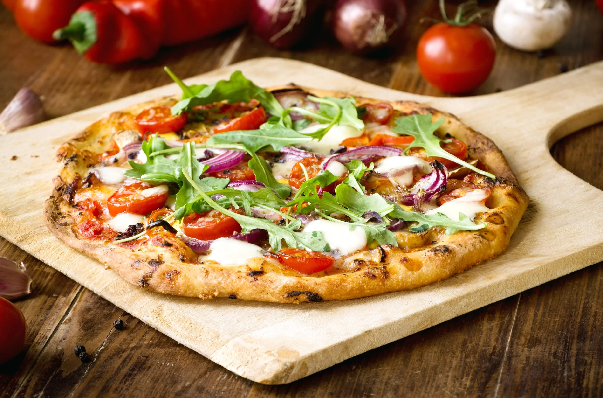 Pizza with cherry tomatoes, arugula, onions, and cheese on a wooden cutting board.