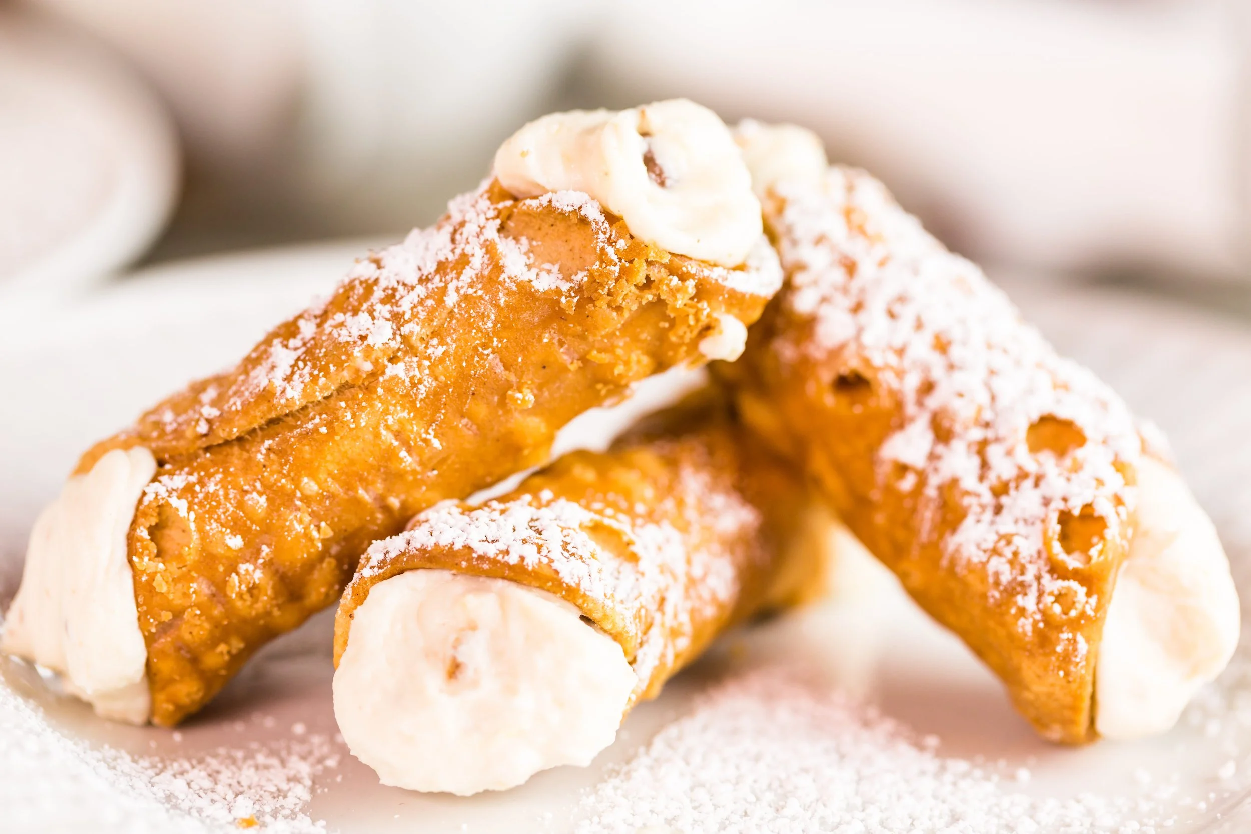 Close-up of cannoli with crispy shell filled with creamy ricotta cheese, topped with powdered sugar and small pieces of chocolate, on a white surface.