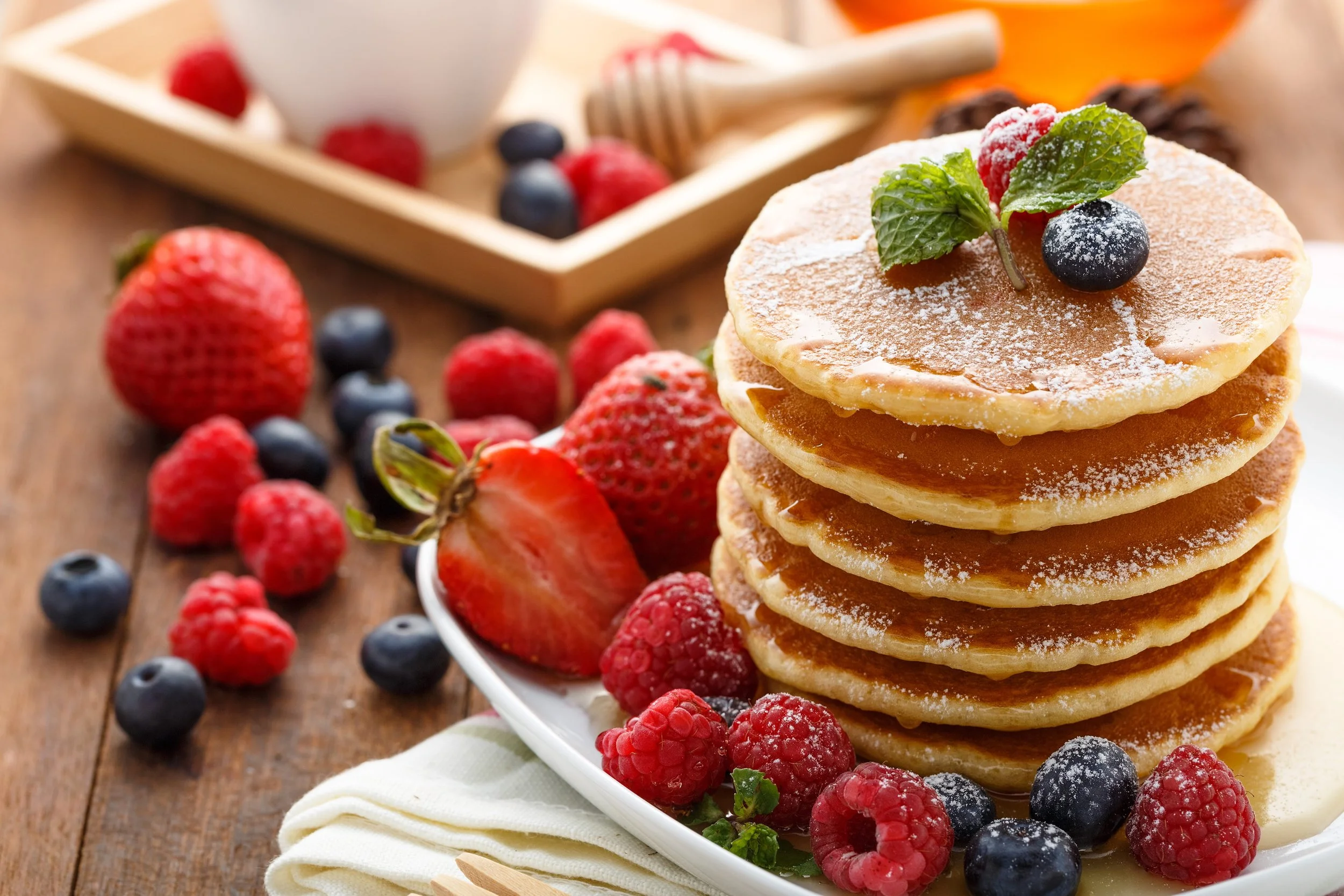 Stack of five golden brown pancakes on a white dish, topped with powdered sugar, raspberries, blueberries, and a sprig of mint, with more berries scattered around on a wooden table.