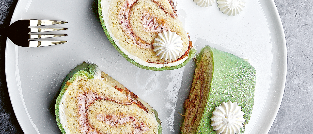 Slices of Swiss roll cake with green sponge, pink filling, and white frosting rosettes on a white plate.