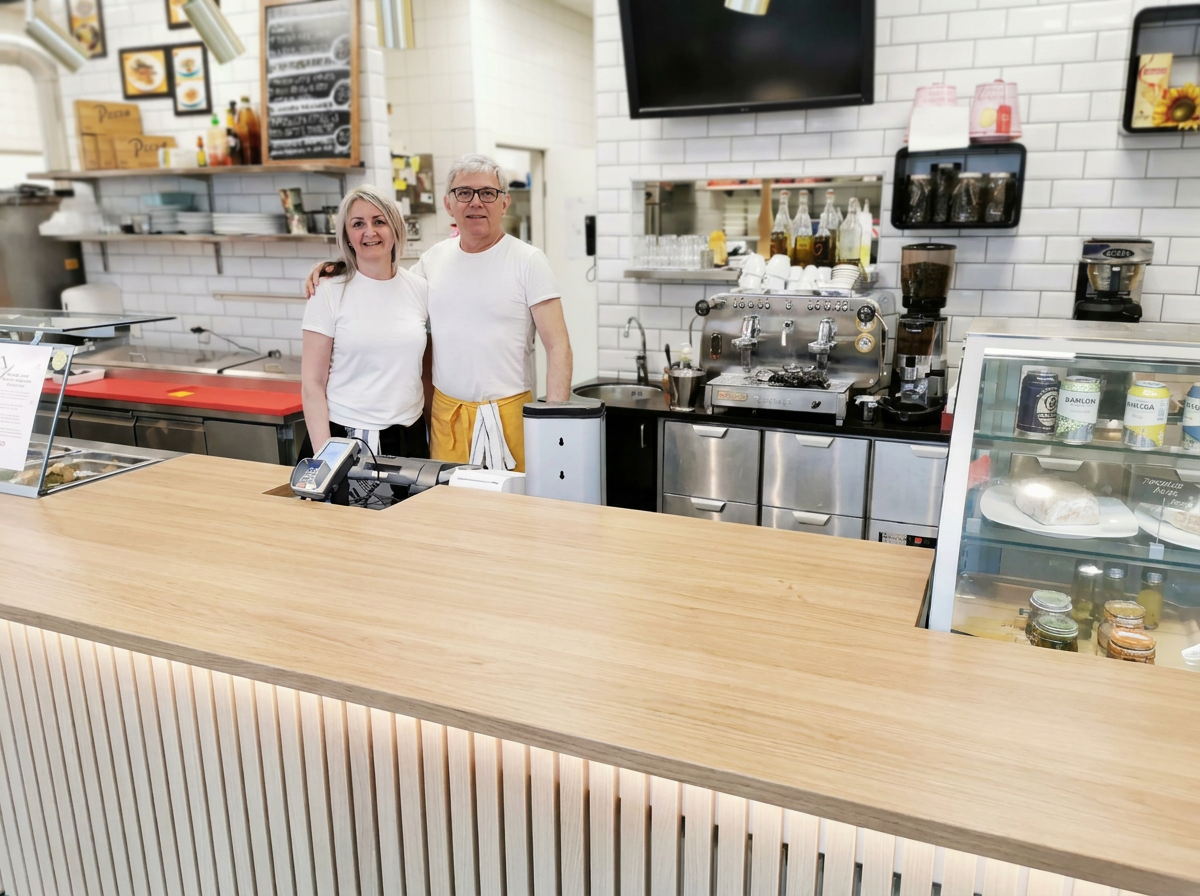 Two people smiling behind a wooden counter in a cafe, with espresso machines and shelves of coffee supplies in the background.