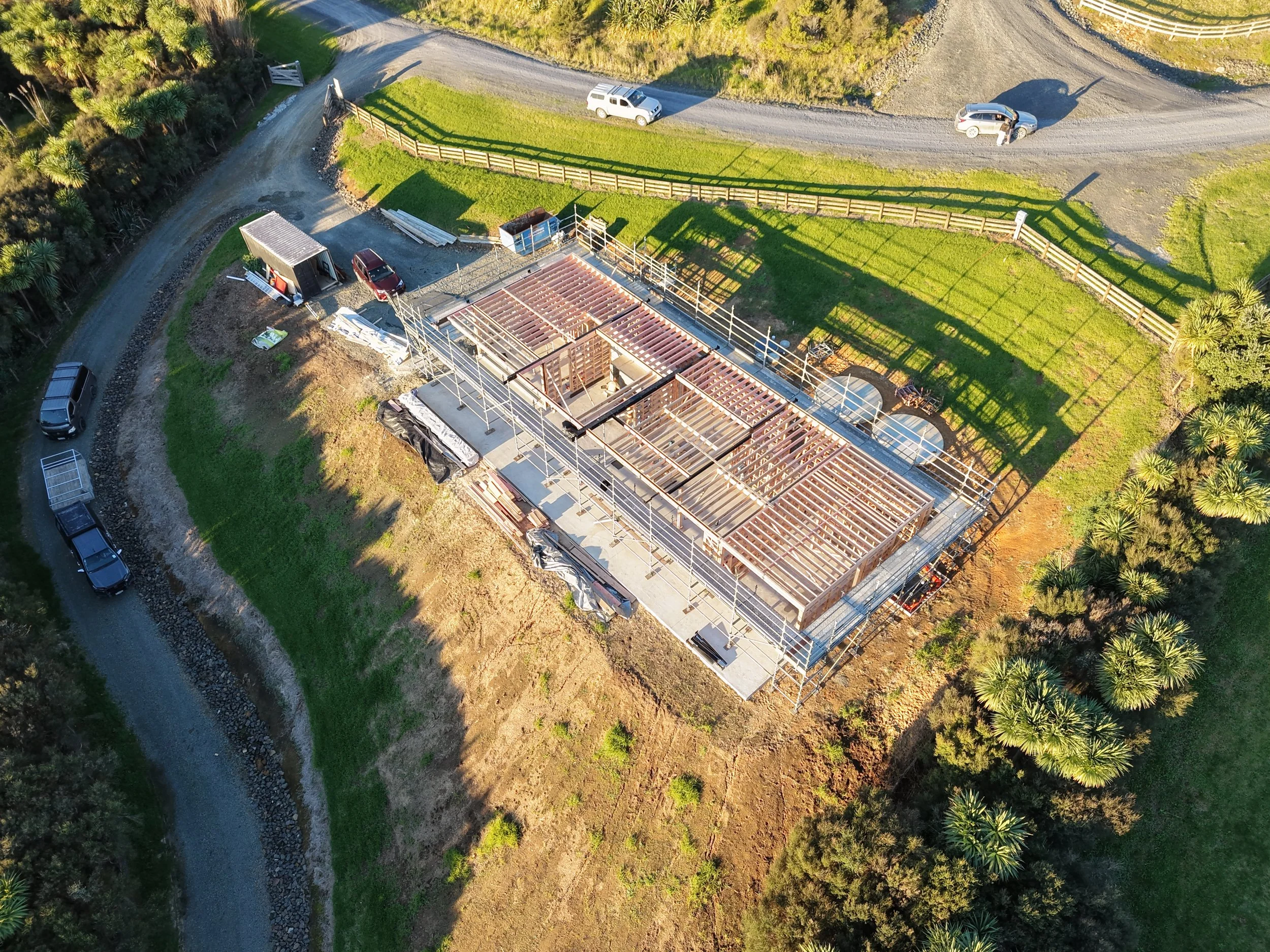 Aerial view of a construction site with a partially built wooden structure, surrounded by green grass, trees, and dirt roads. Several vehicles are parked around the site.