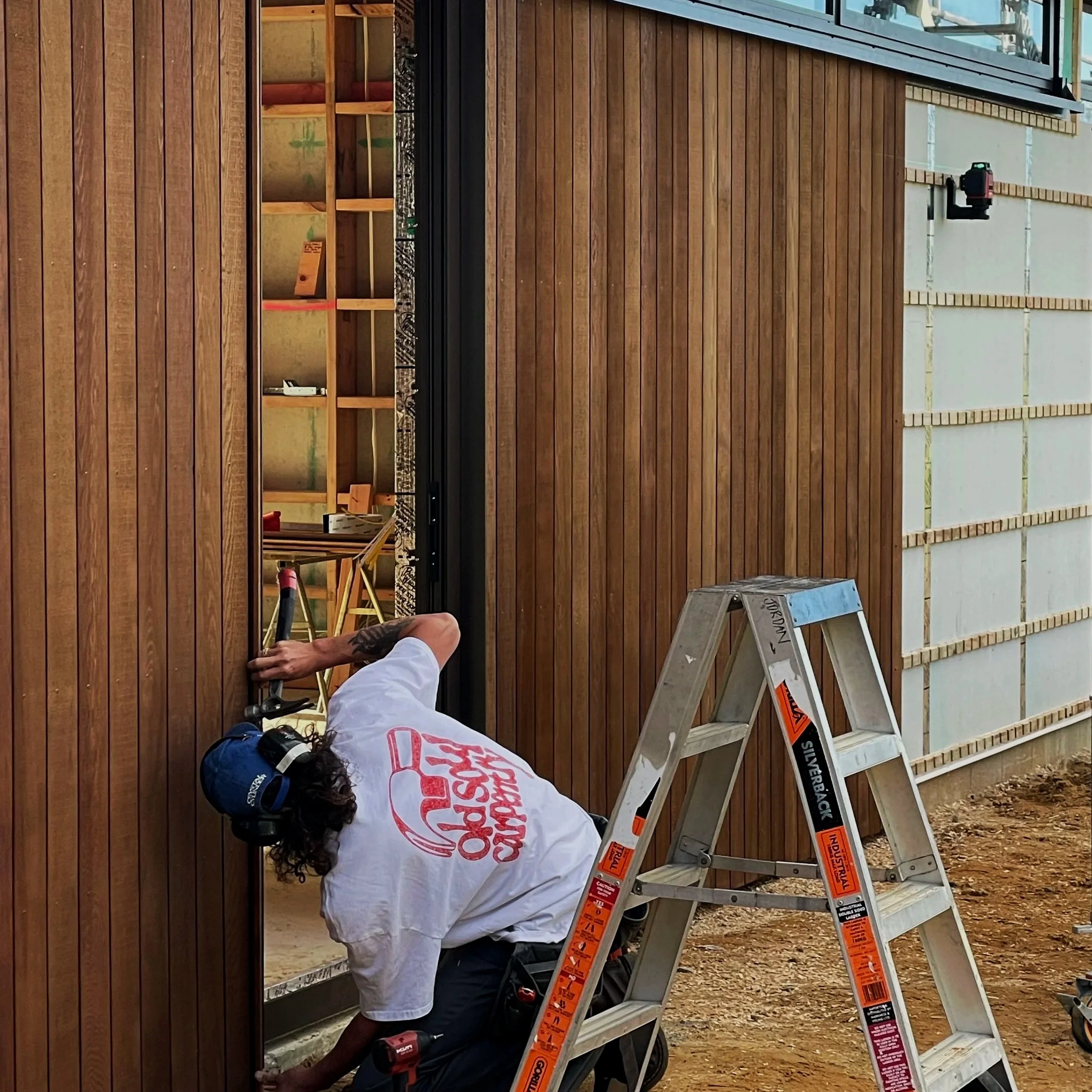 A person working on a a cedar home  standing on a ladder, wearing a white shirt, dark pants, and a hat, with construction tools nearby.
