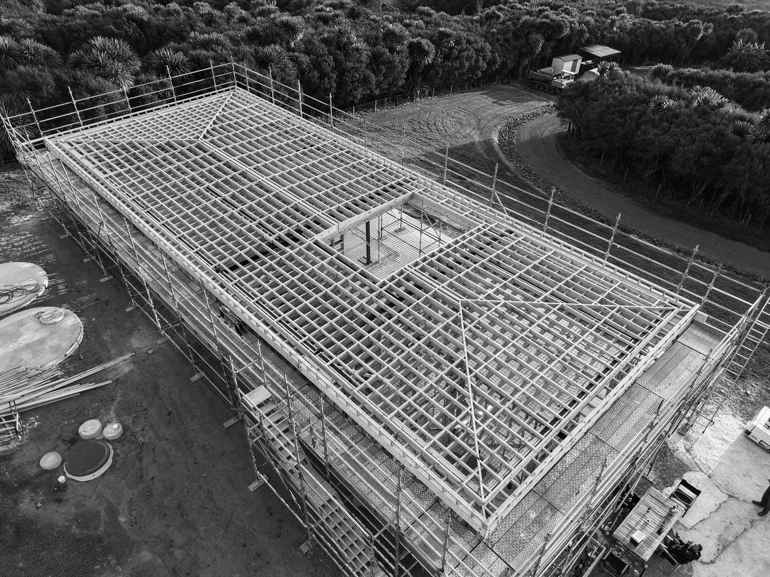 Black and white aerial view of a building under construction with a wooden framework, surrounded by scaffolding, with trees and a small train in the background.