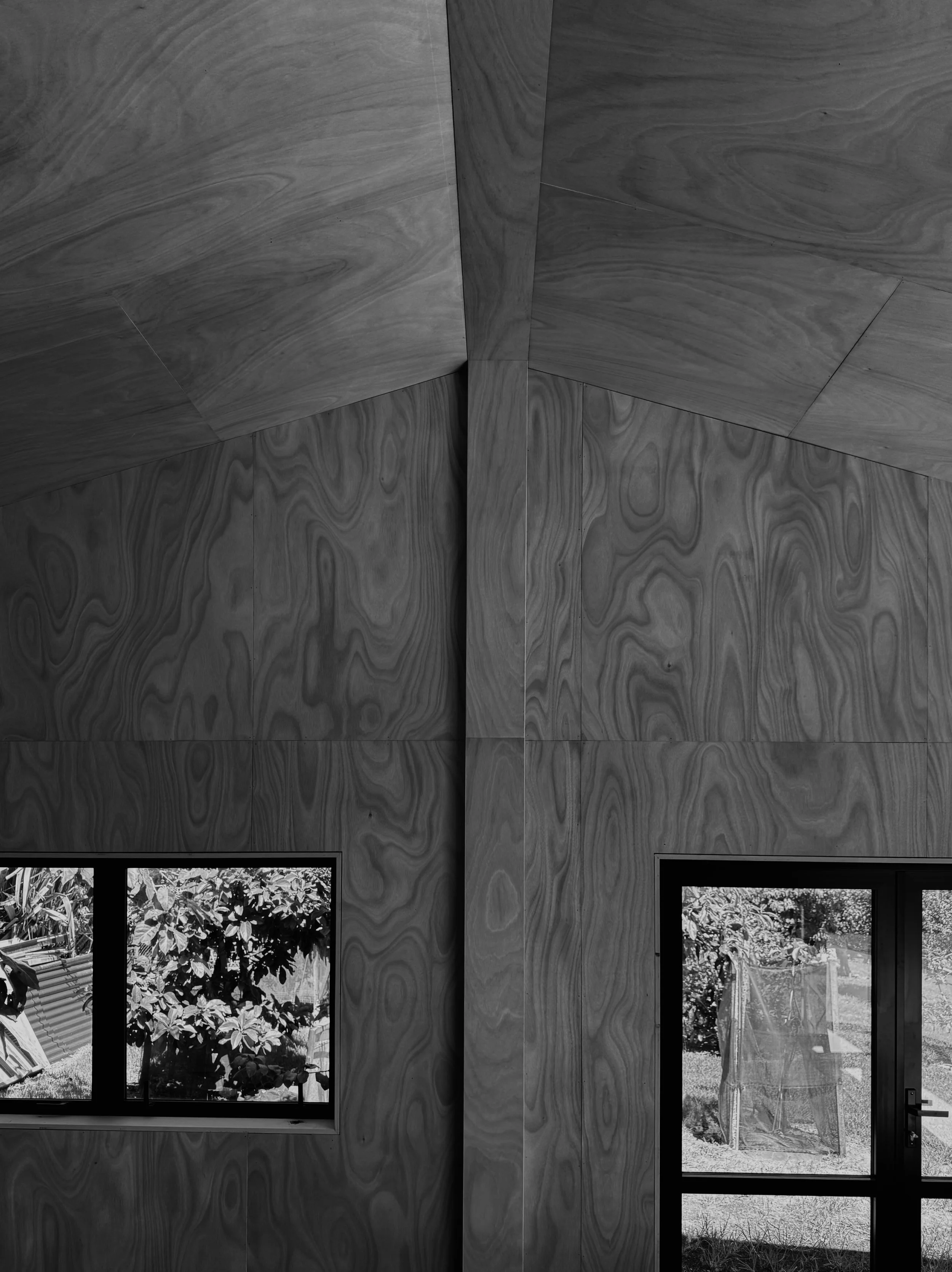 Black and white photograph of a room with a wooden wall, two windows showing trees outside, and a partial view of a garden or outdoor area.