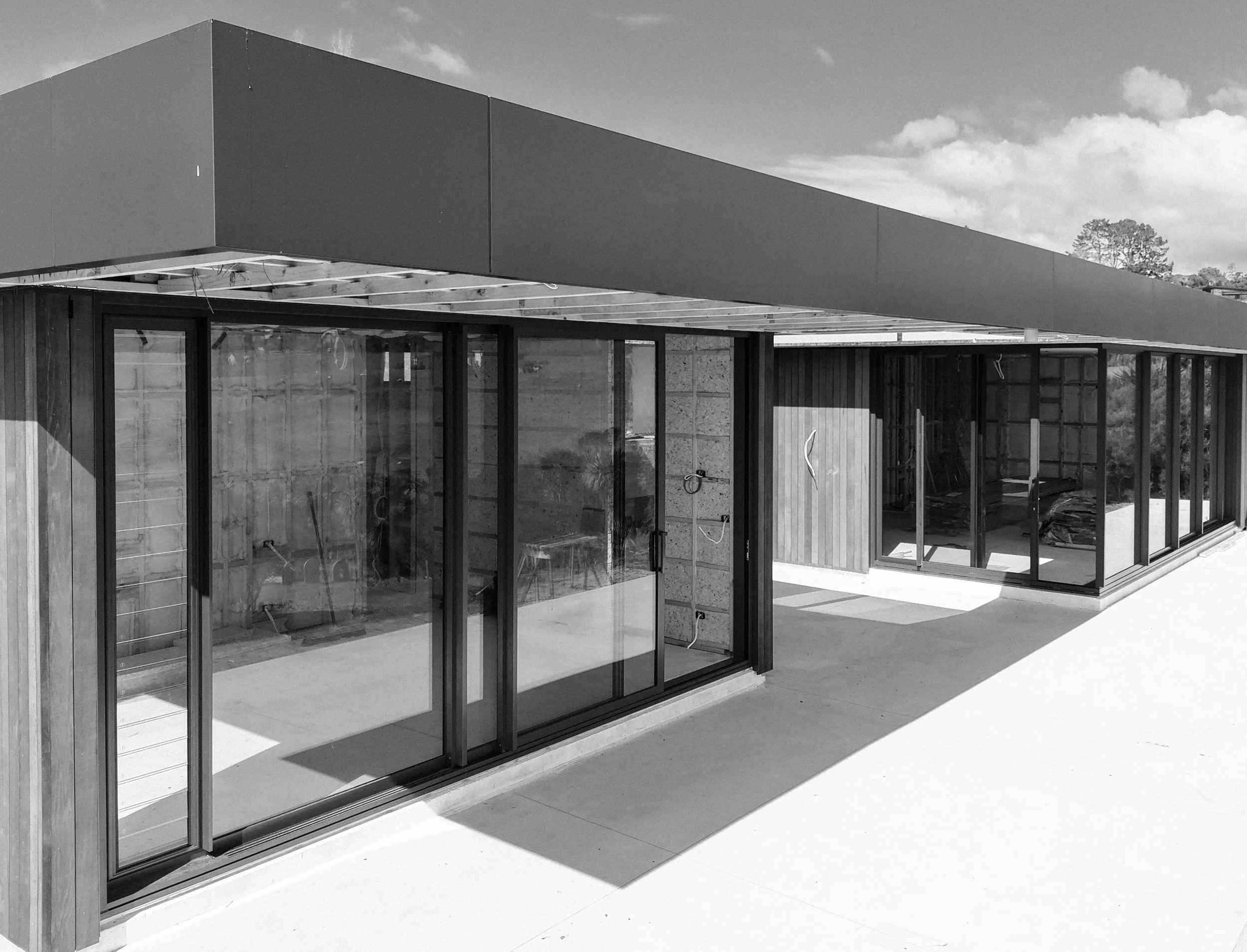 Modern building under construction with large glass windows and a flat roof, construction materials visible, and trees in the background under a partly cloudy sky.