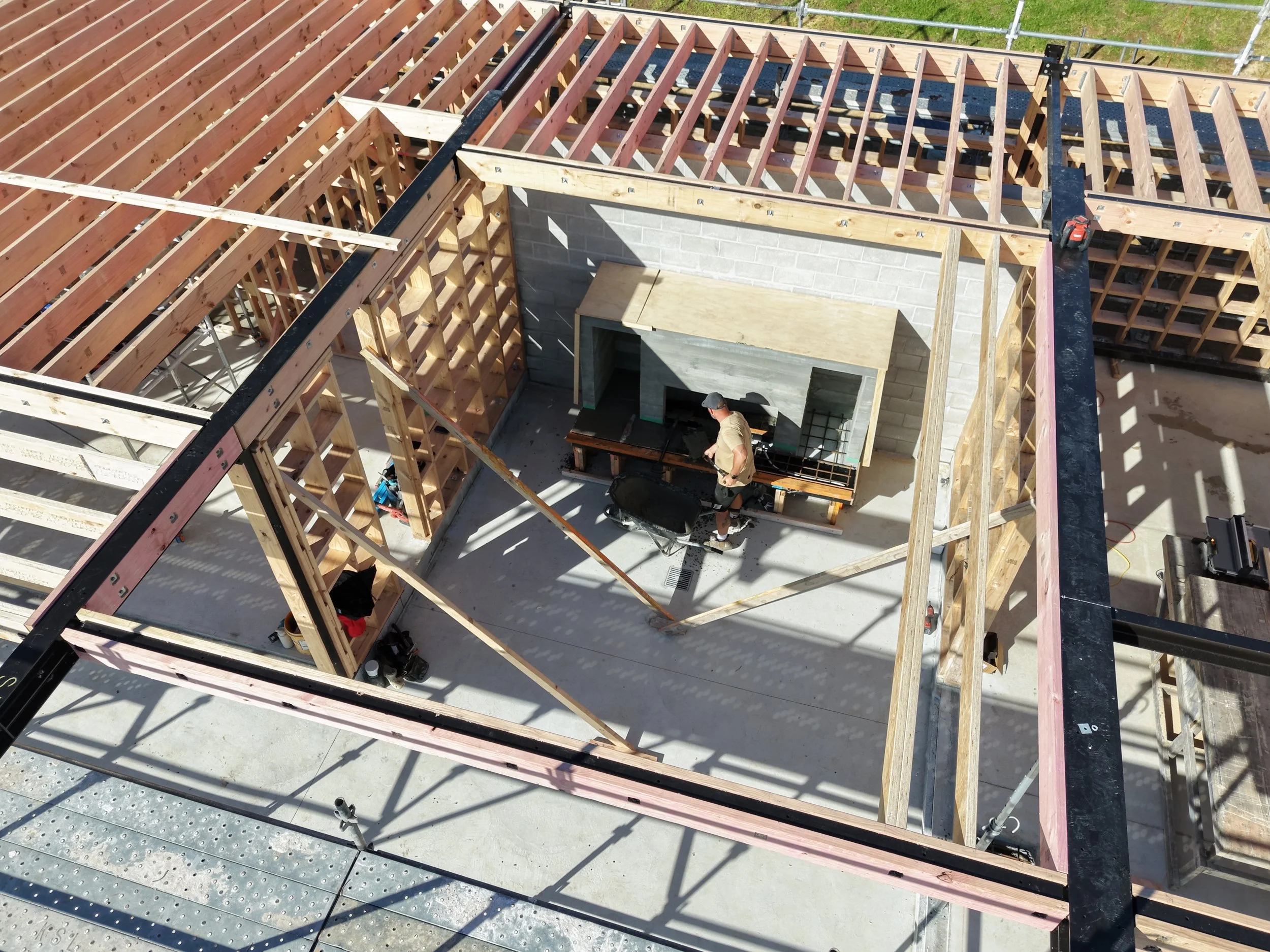 Construction site with wooden framing and a person working in the area. The structure appears to be in the early stages of building, with scaffolding and building materials visible.
