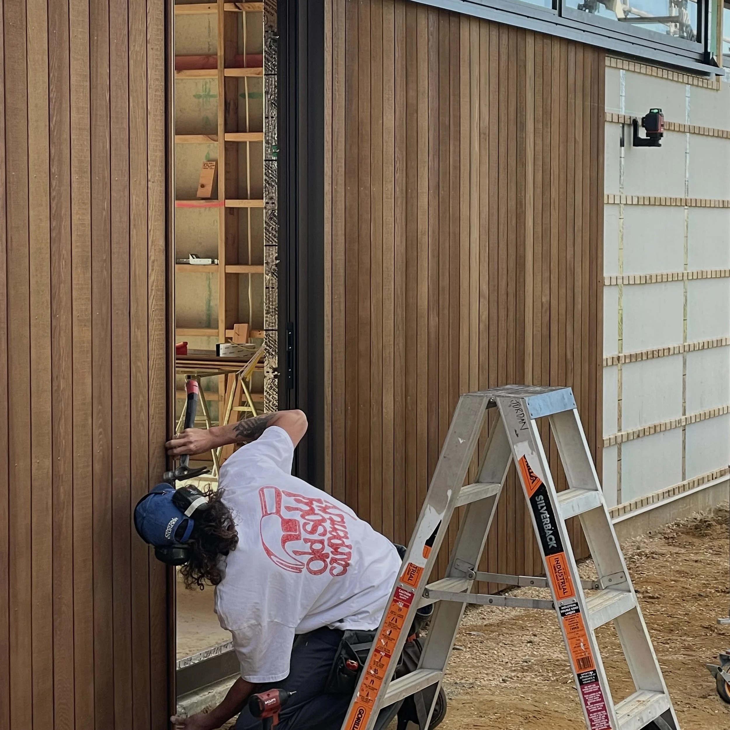 Construction worker installing wooden siding on a building, using a ladder and power tools, with some unfinished sections of the wall visible.