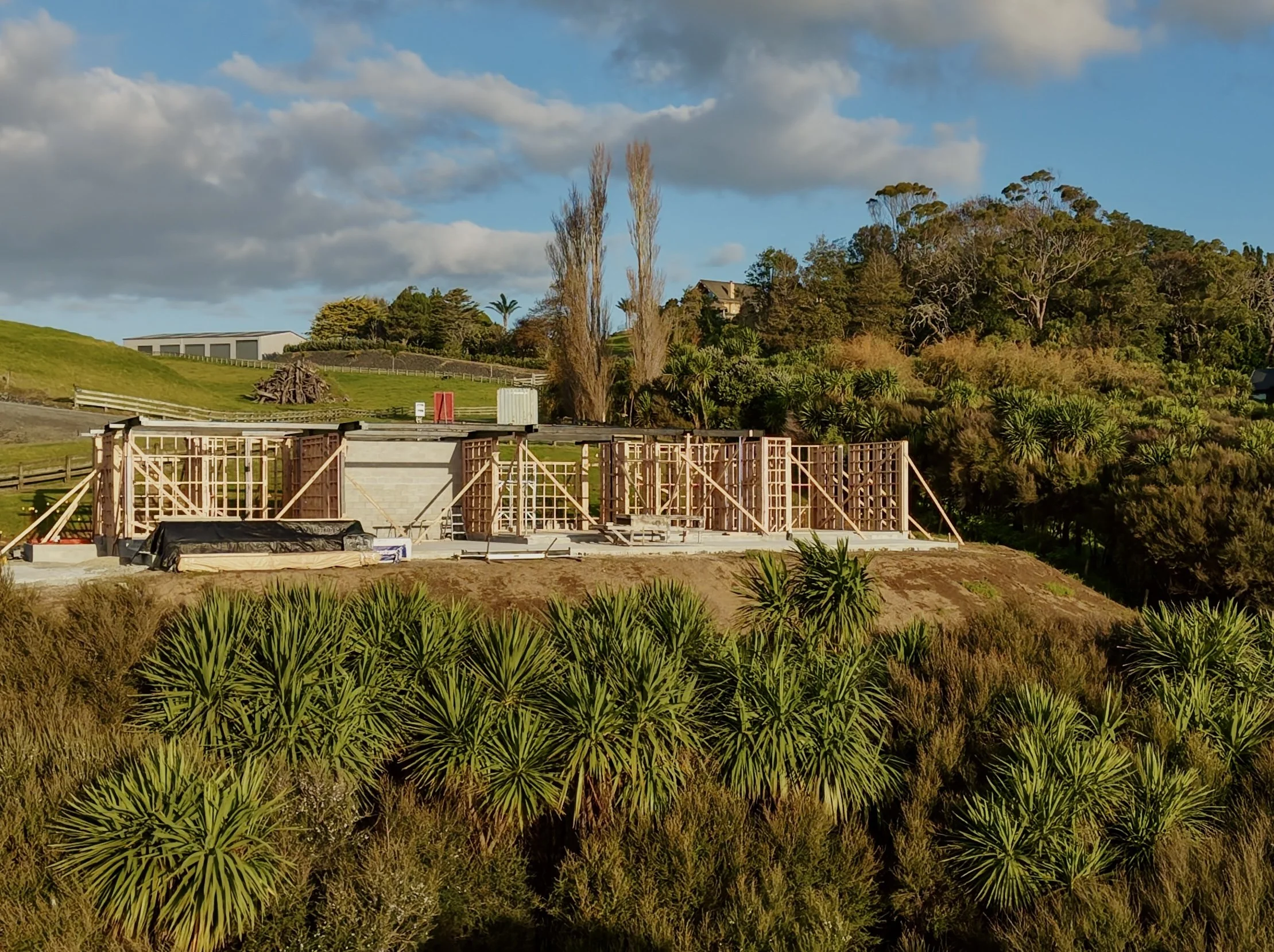 Under construction site with wooden framing, surrounded by green shrubbery and trees, under a partly cloudy sky.