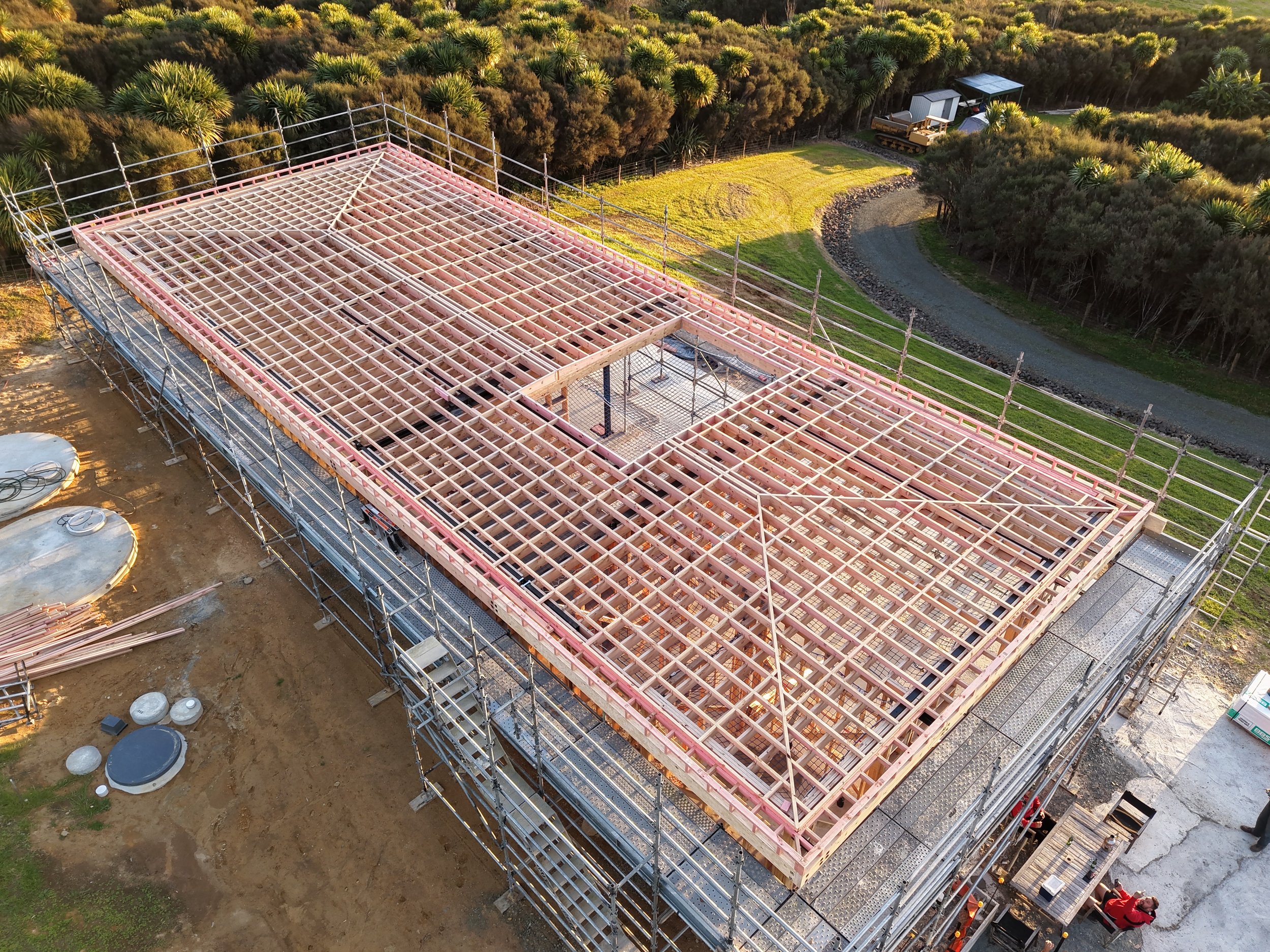 Construction site with wooden framework for a building, surrounded by scaffolding, in a green area with trees and a gravel path.