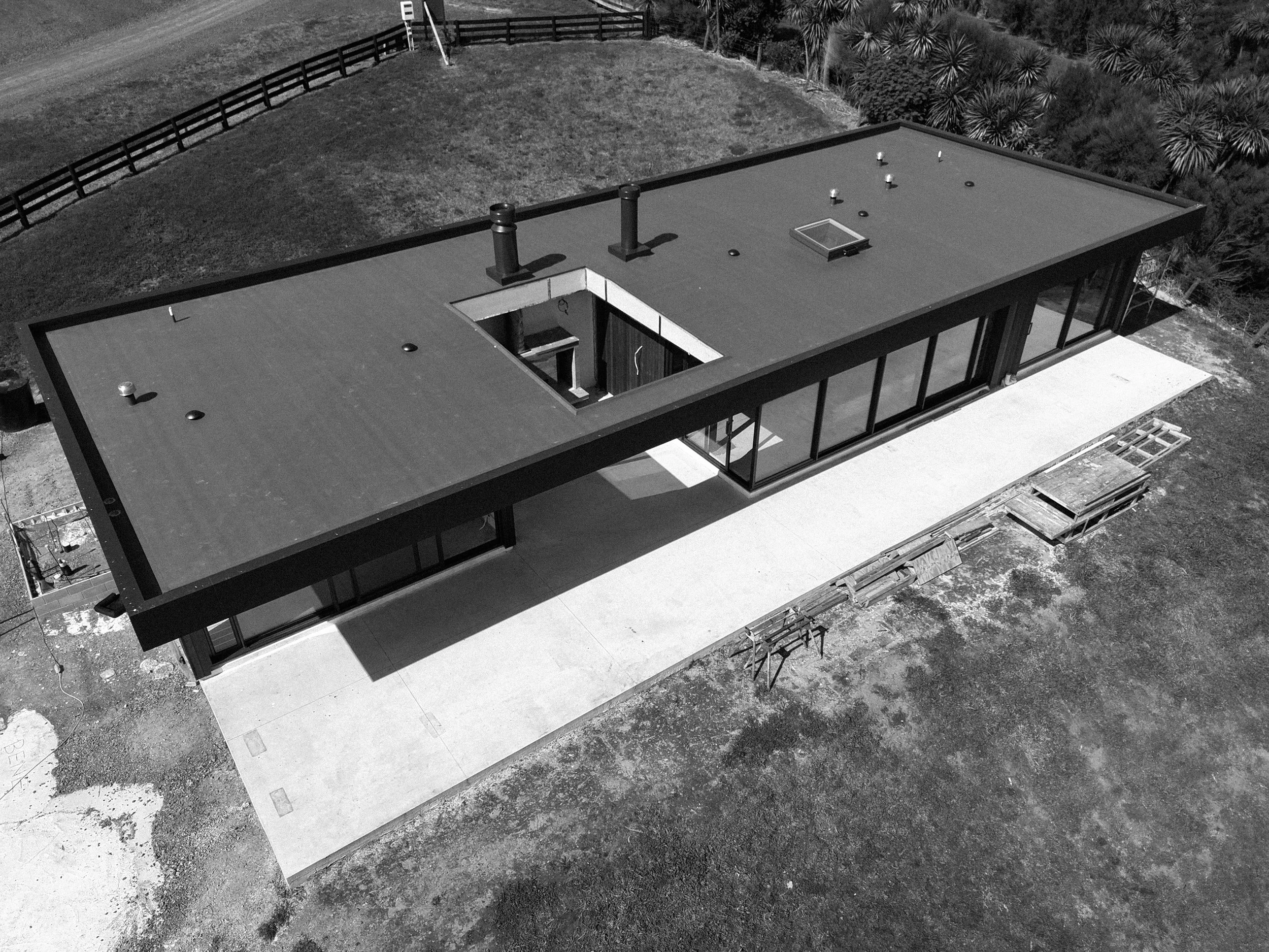 A modern flat-roof house under construction with black pipes and vents on the roof, located in a grassy area with trees and a wooden fence nearby.