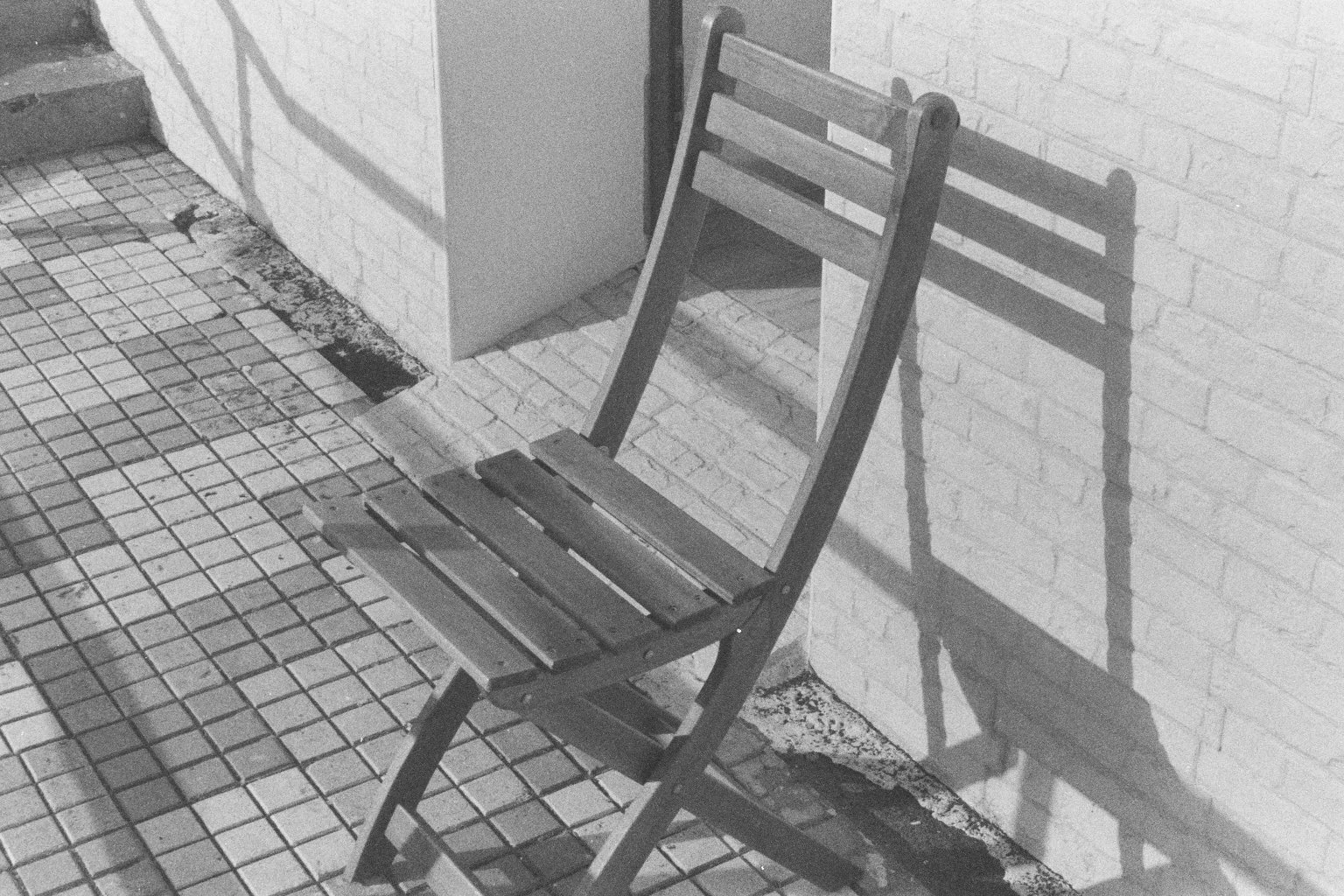 Ein unbequemer Holzstuhl steht auf einer Gehwegfliese vor einer Ziegelwand, seine Schatten sind sichtbar auf der Wand