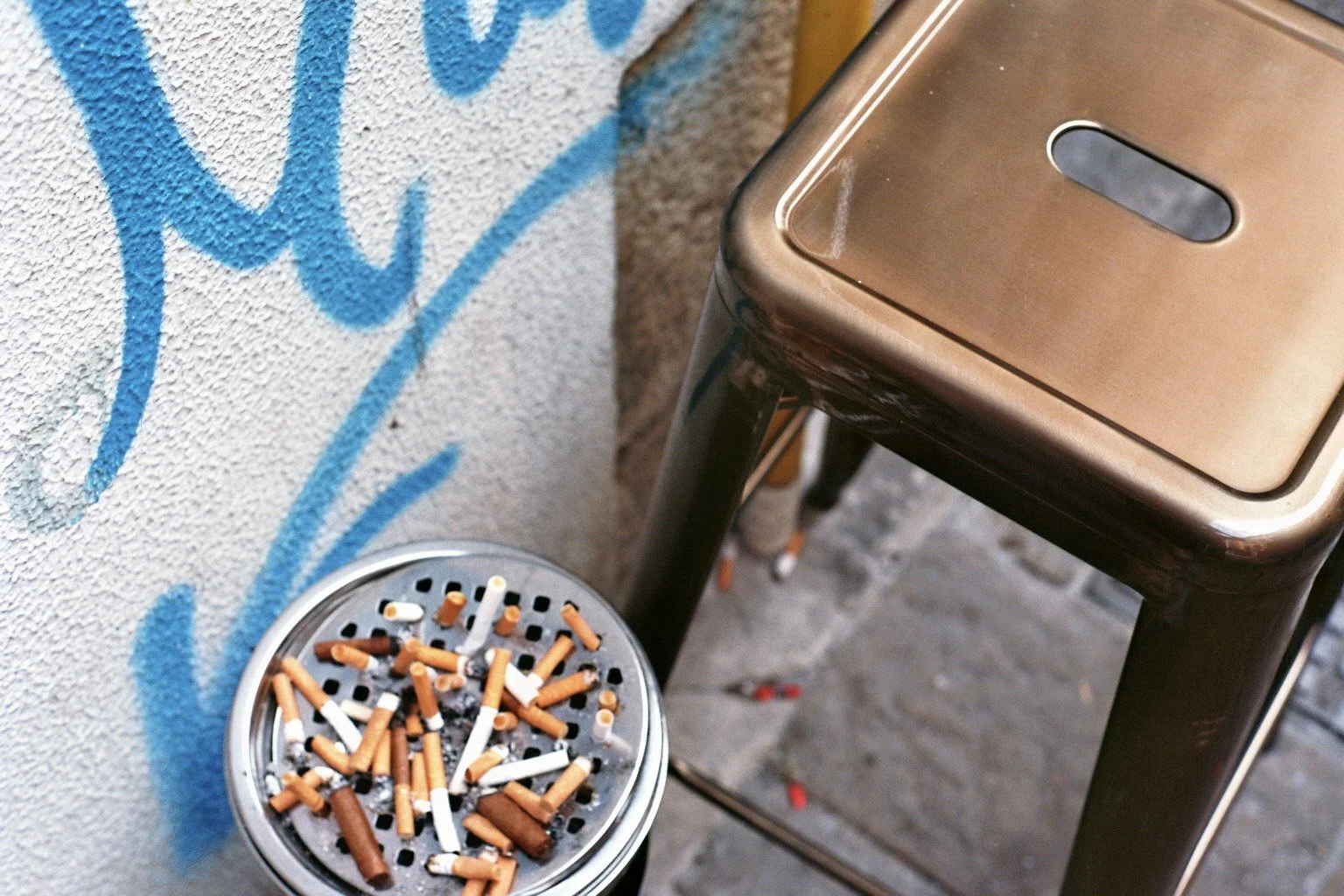 Aschenbecher mit vielen Zigarettenkippen neben einem braunen Mülleimer auf einem grauen Boden, an einer Wand mit blauer Graffiti. 