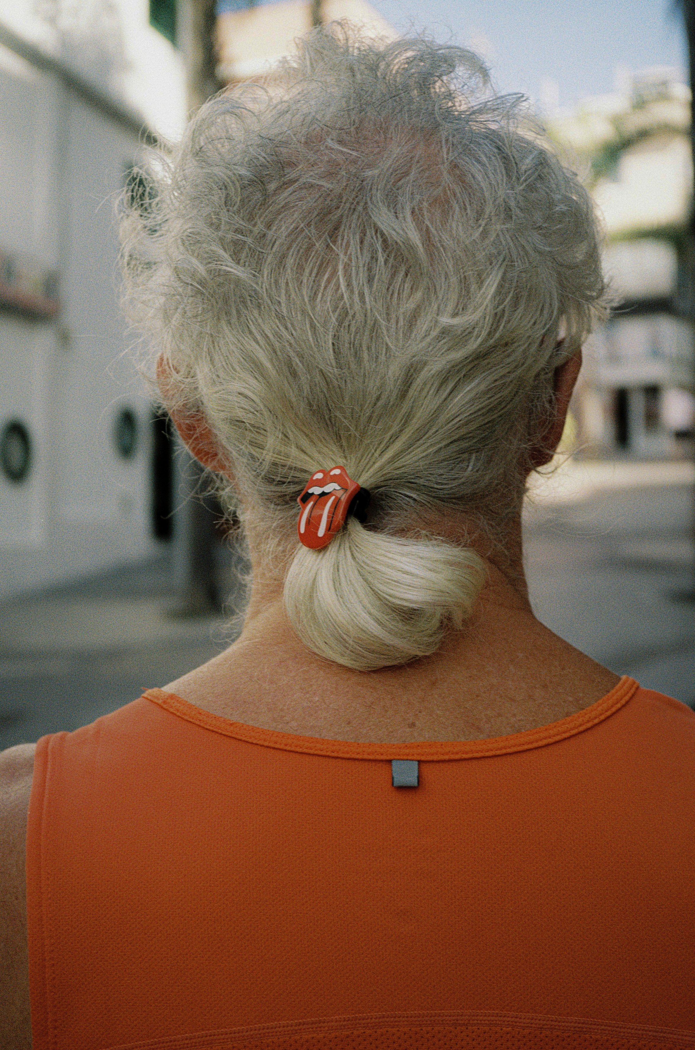Nahaufnahme des Rückens eines älteren Menschen mit grauem, lockigem Haar, das zu einem Zopf gebunden ist, und einem orangefarbenen Oberteil, draußen mit Gebäuden im Hintergrund.