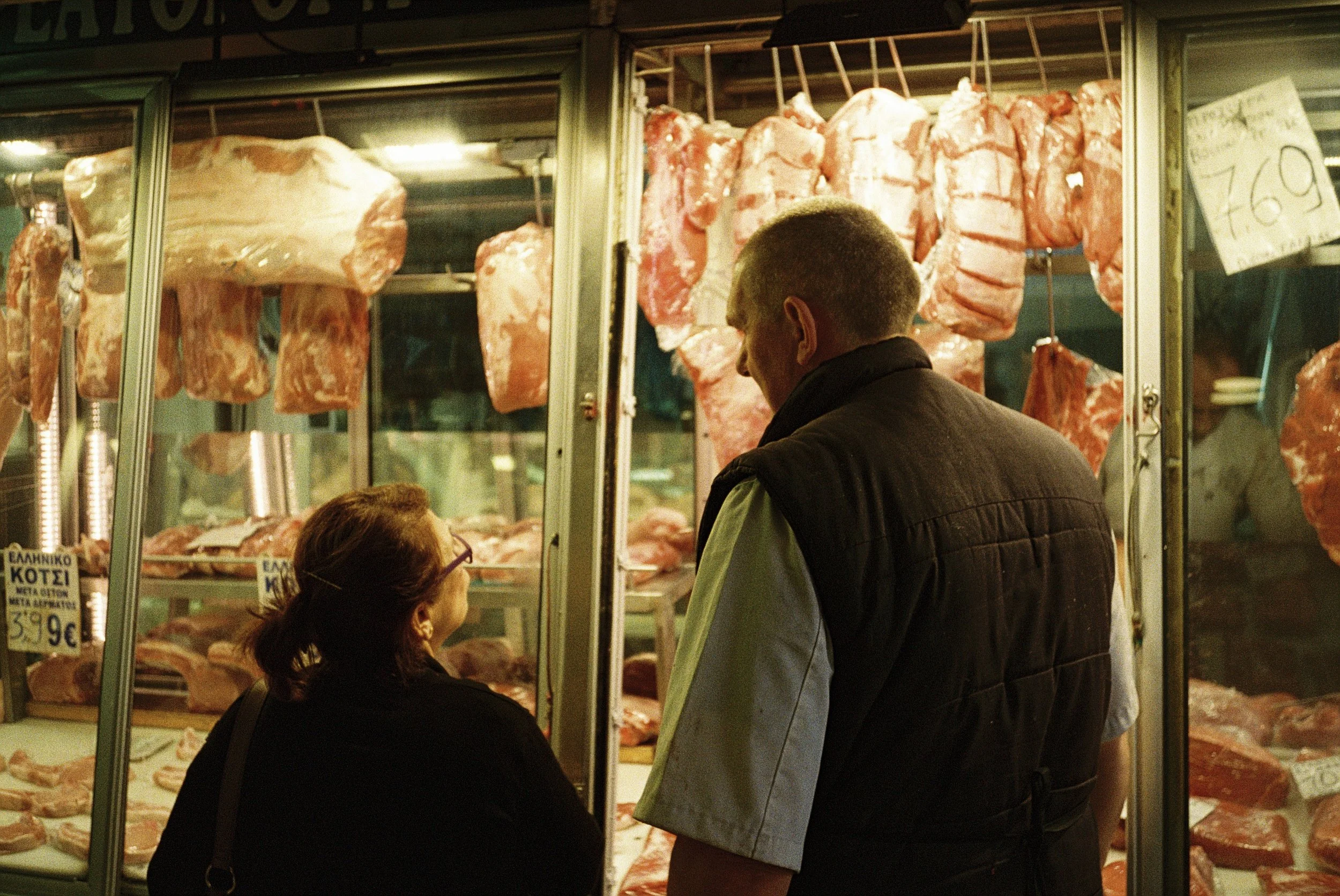 Menschen beim Fleischkauf an einem Fleischstand auf einem Markt, mit Fleischstücken im Kühlregal im Hintergrund.