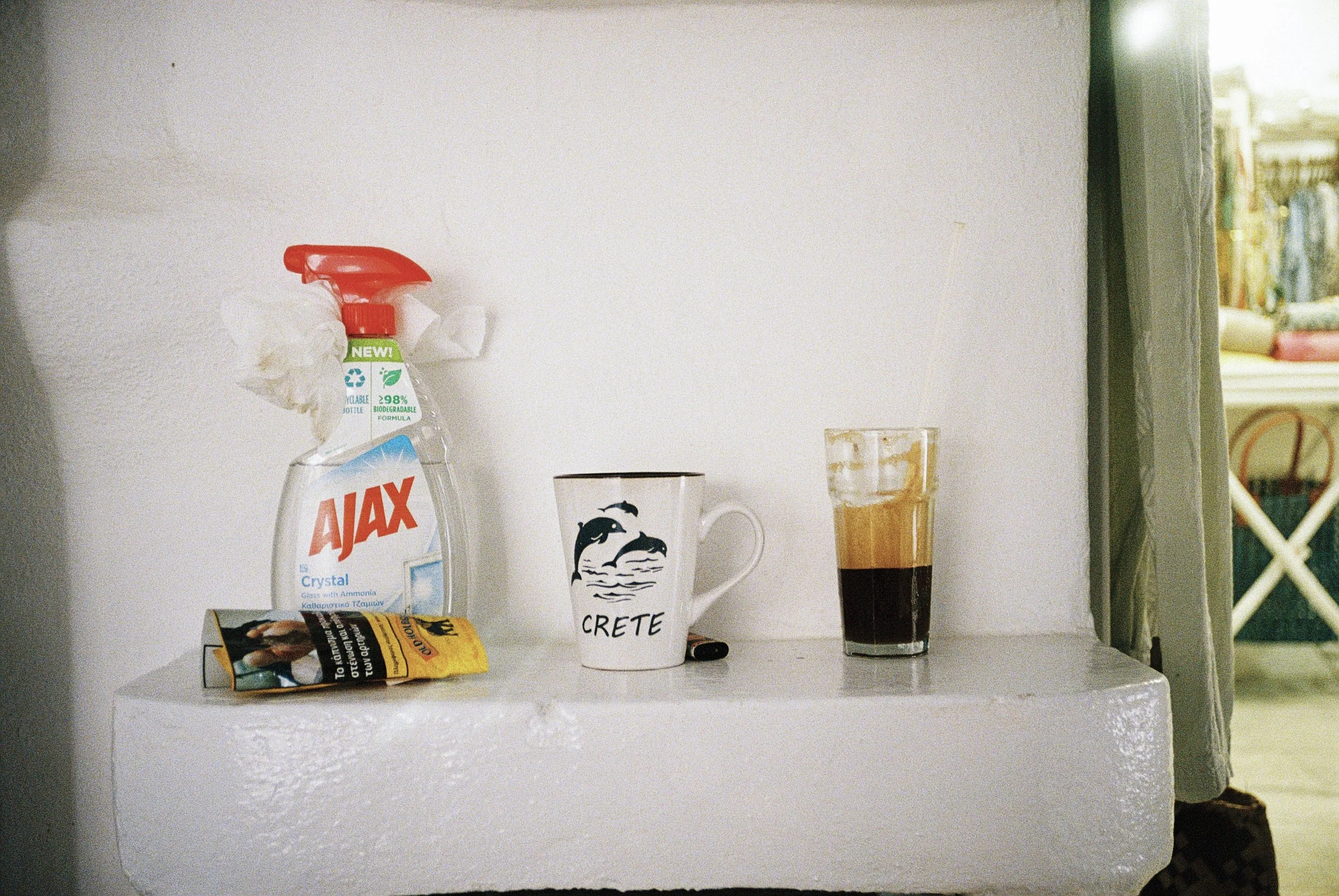 Auf einem weißen Tisch befindet sich eine Tasse mit dem Wort "CRETE" und einem Delfin-Design, ein Glas mit dunkler Flüssigkeit, eine Flasche von "Ajax" Reinigungsmittel, ein müllbätzchen und ein Packung mit Chips. Im Hintergrund sind Regale mit Büche