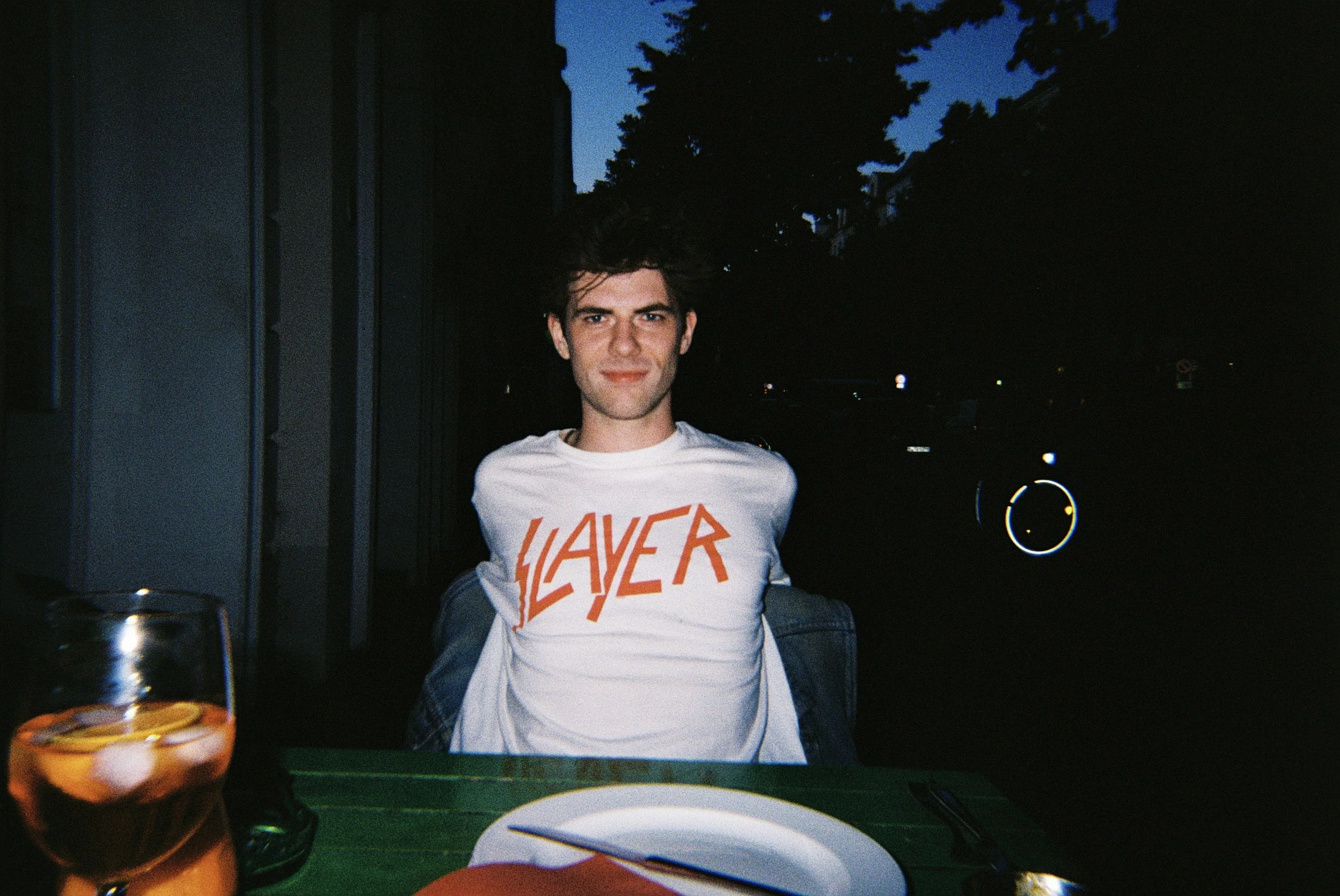 Ein junger Mann sitzt abends an einem Tisch im Freien, trägt ein weißes T-Shirt mit roten Buchstaben und lächelt in die Kamera. Im Vordergrund sind eine orangefarbene Flasche und ein Teller zu sehen.