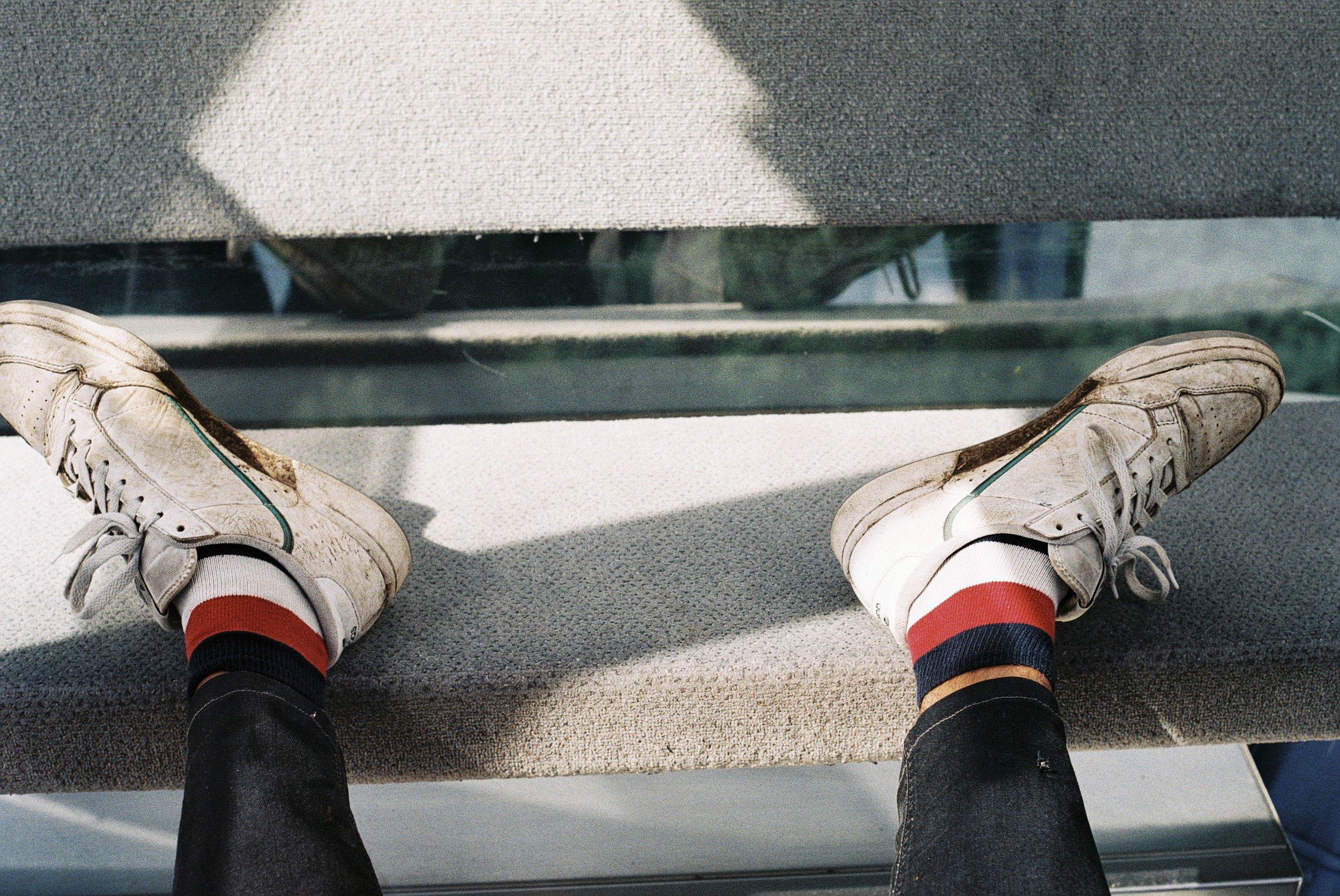 Perspektive eines Sitzenden mit weißen Schuhen, roten und schwarzen Socken, Blick auf den Boden und Füße vor einer Glastrennwand mit Blick nach unten.