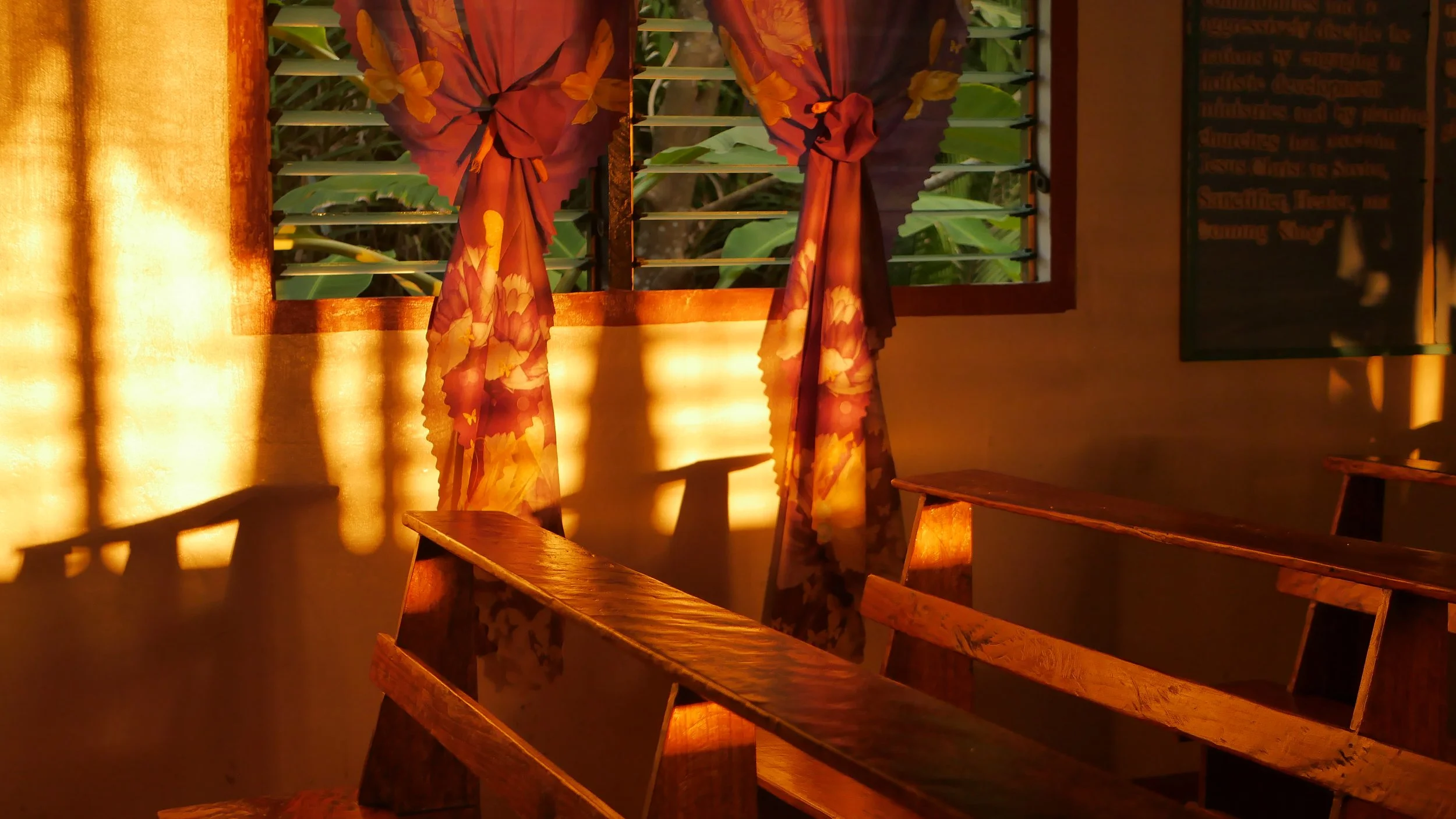 Ein Raum mit warmem Licht, Holzbänken, bunten Vorhängen an einem Fenster und einer Tafel auf der rechten Seite.
