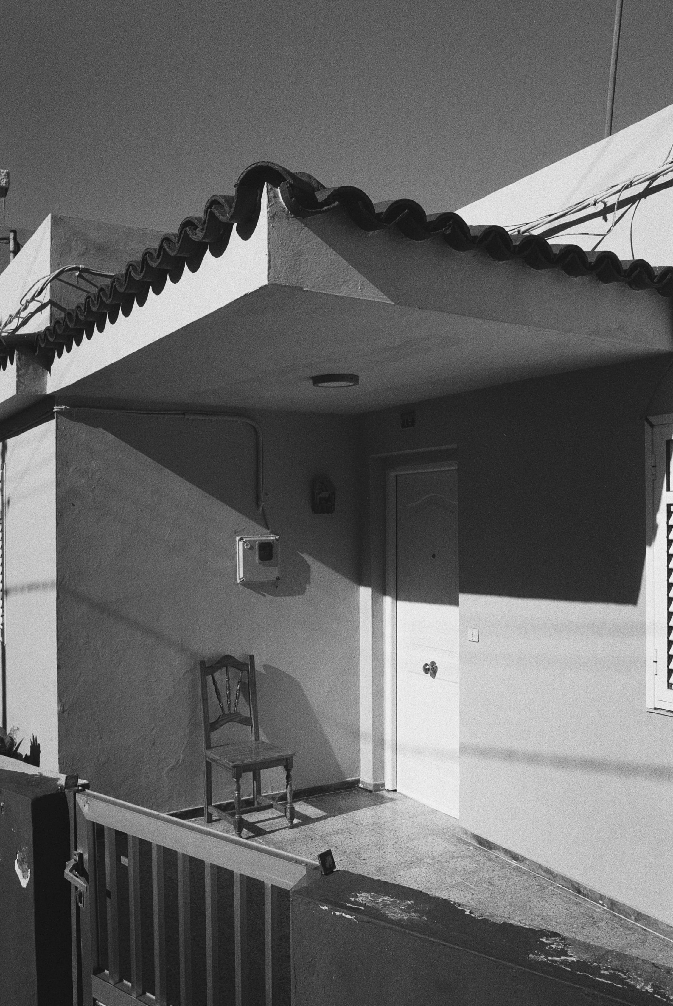 Das Bild zeigt die Ecke eines Gebäudes mit einem weißen Balkon, terrakottafarbigem Dach und einer weißen Tür. Es gibt eine einfache Holzstuhlsitzgelegenheit, Kabel und Schatten.