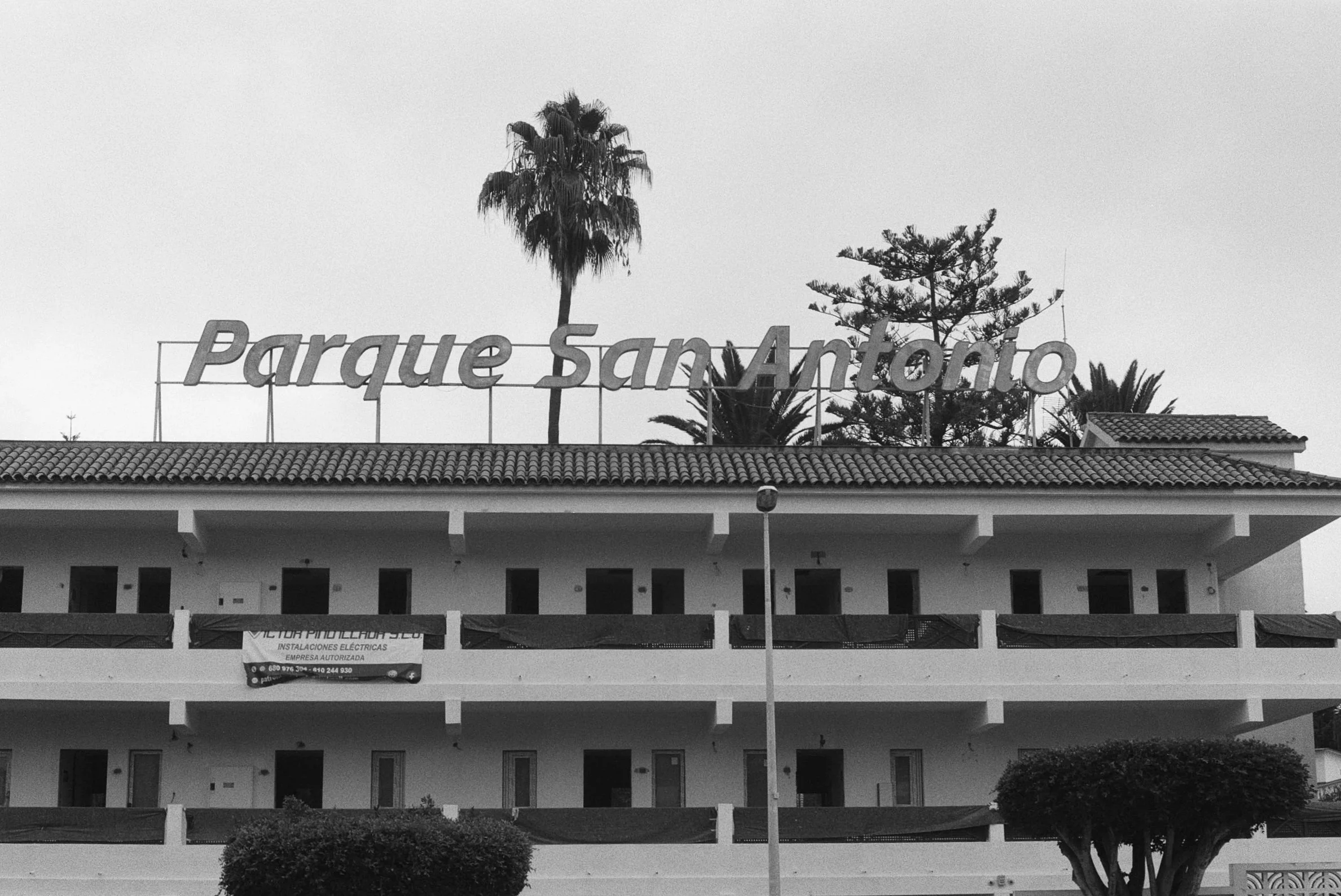 Ein Gebäude mit einem großen Schild 'Parque San Antonio' auf dem Dach, umgeben von Palmen und Kiefern, im Hintergrund ein grauer Himmel.