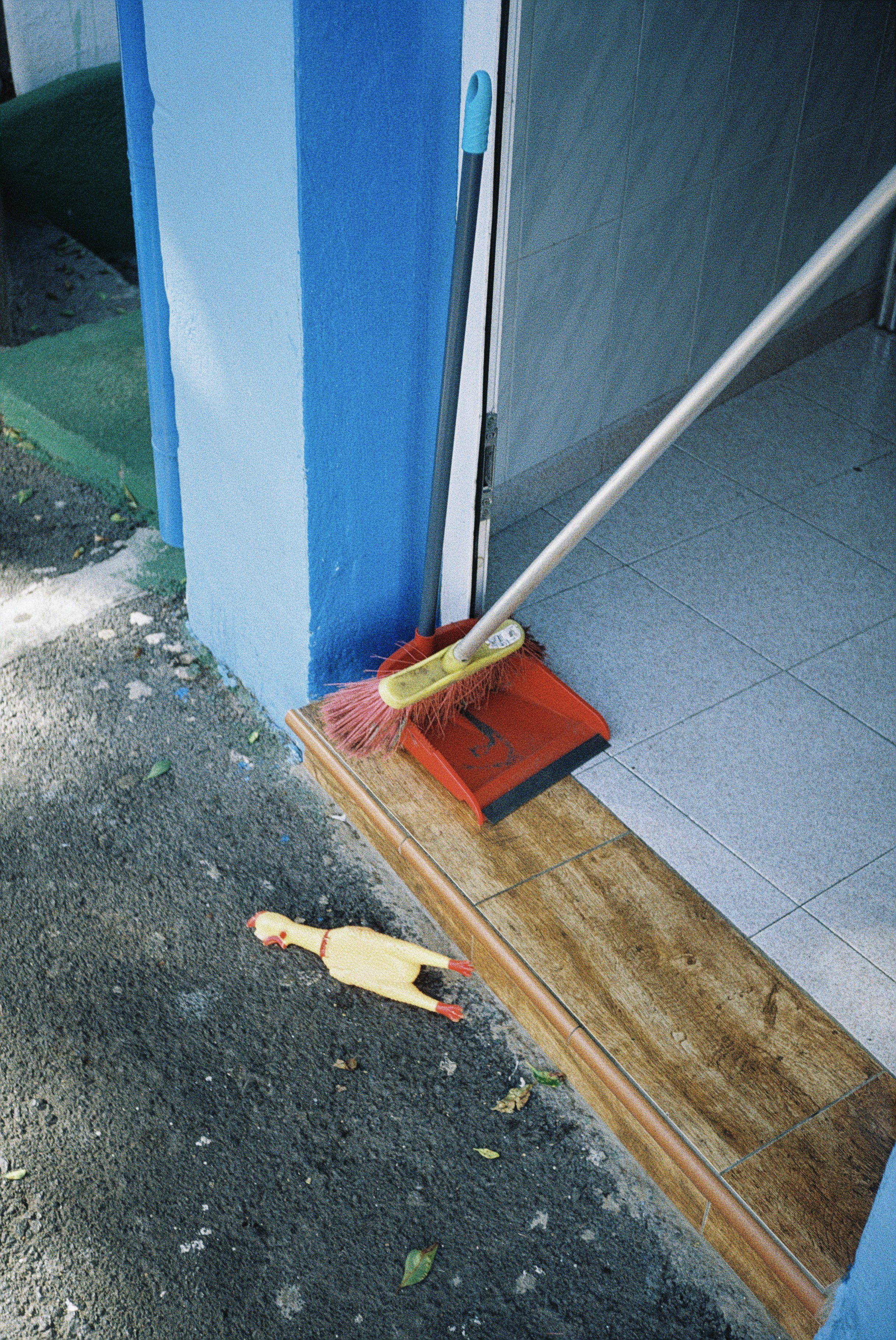 Straße mit Reinigungsgeräte, darunter ein Besen, eine Kehrschaufel und eine Gummiente auf dem Boden, vor einer Tür mit Fliesen im Hintergrund.