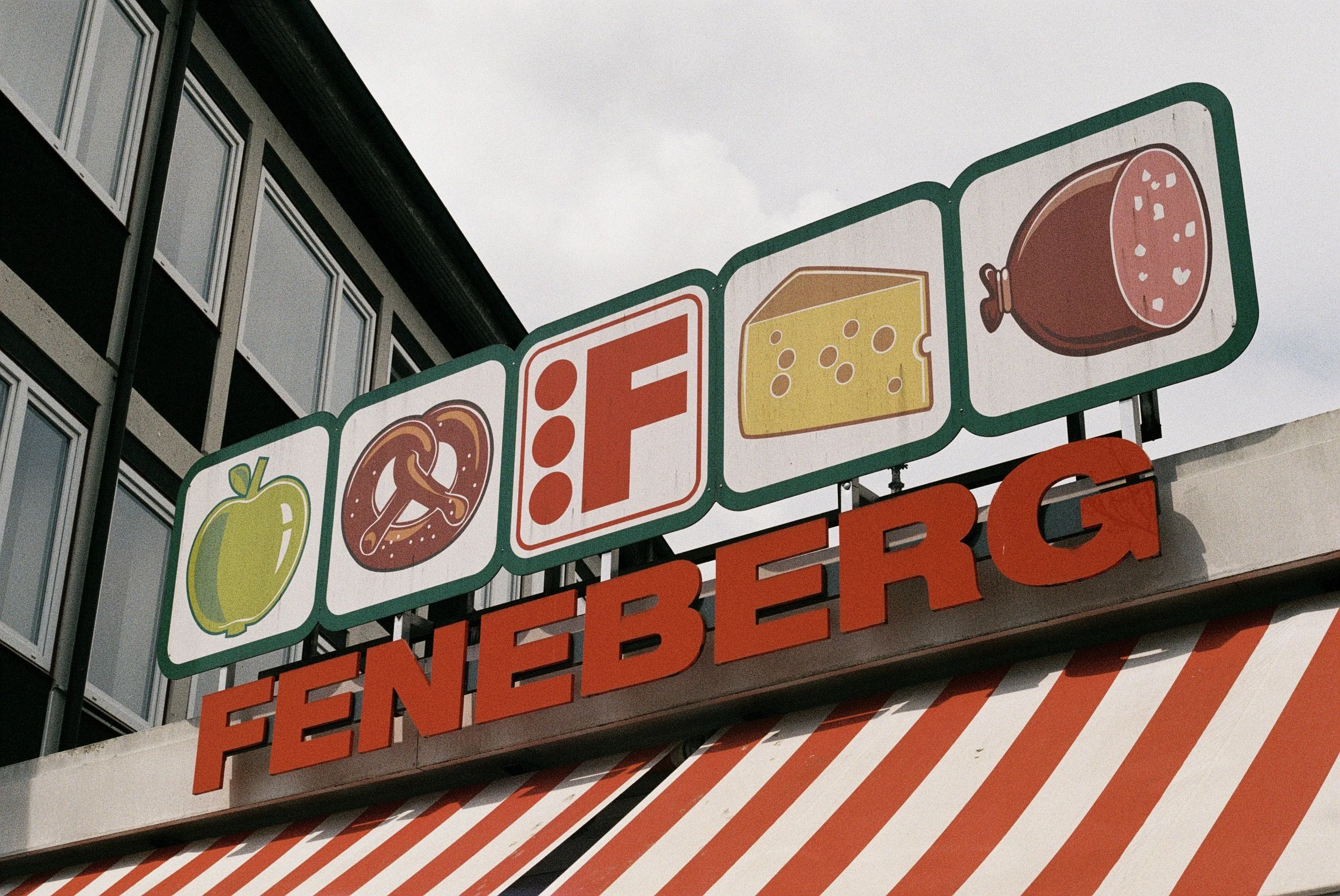 Blick auf ein Werbeschild mit Bildern von Lebensmittelprodukten wie Paprika, Käse, Wurst und Pizza vor einem Mehrfamilienhaus mit mehreren Fenstern.