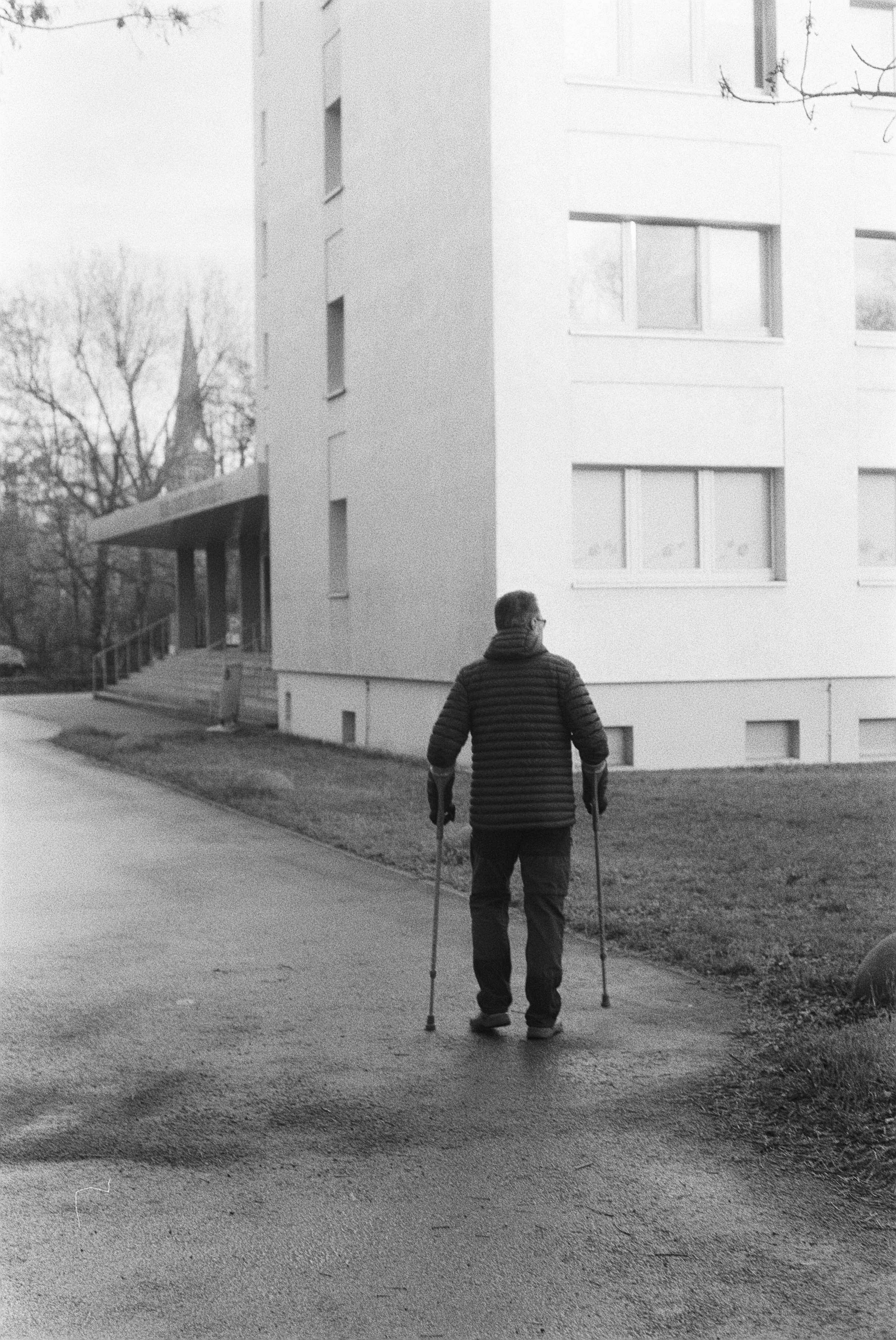 Ein älterer Mann mit Gehhilfen geht auf einer Straße in einer Wohngegend, im Hintergrund sind mehrstöckige weiße Gebäude und Bäume ohne Blätter.