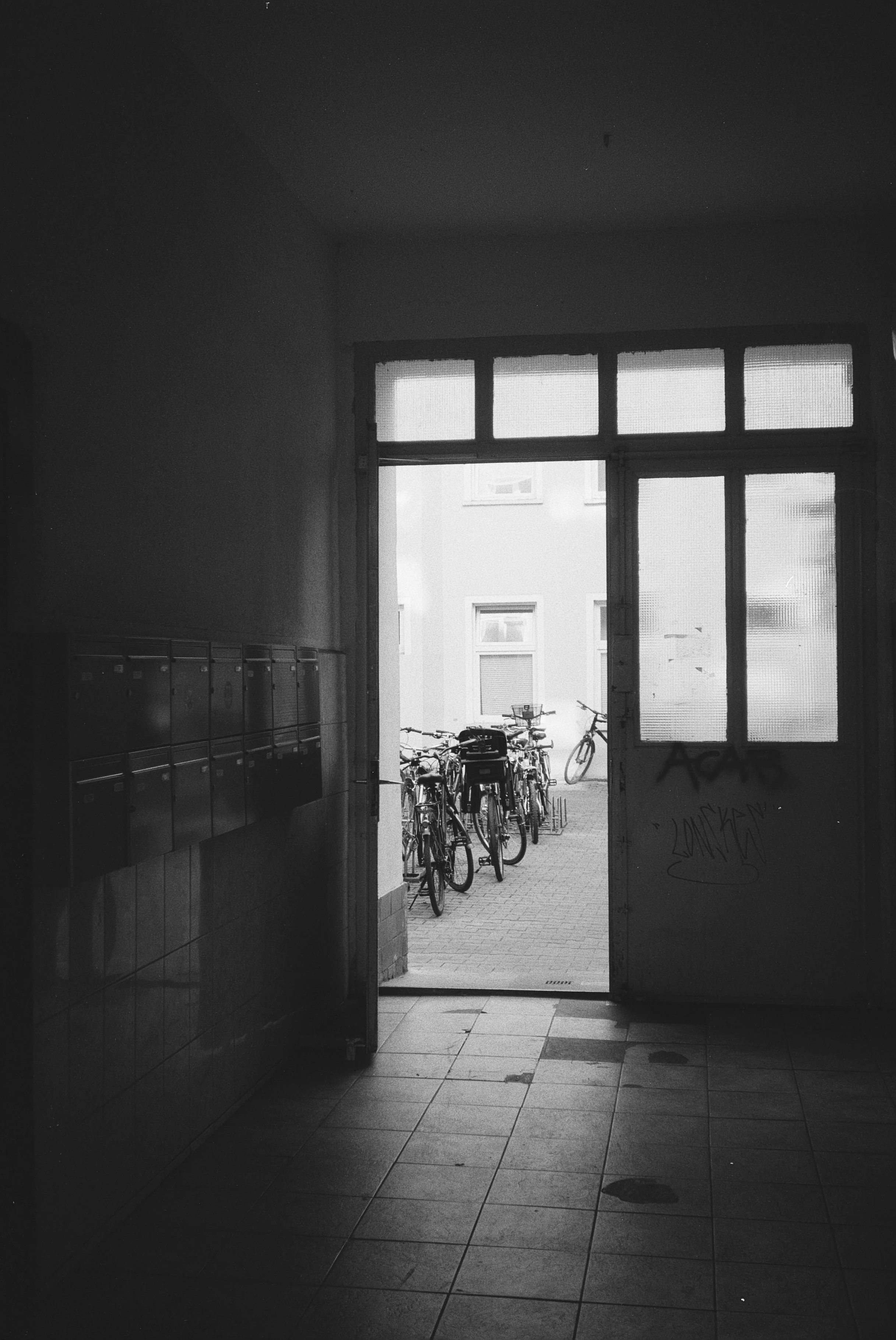 Schwarz-weiß-Fotografie einer Eingangstür zu einem Fahrradkeller, durch den Fahrräder im Freien sichtbar sind, mit Gebäuden im Hintergrund.