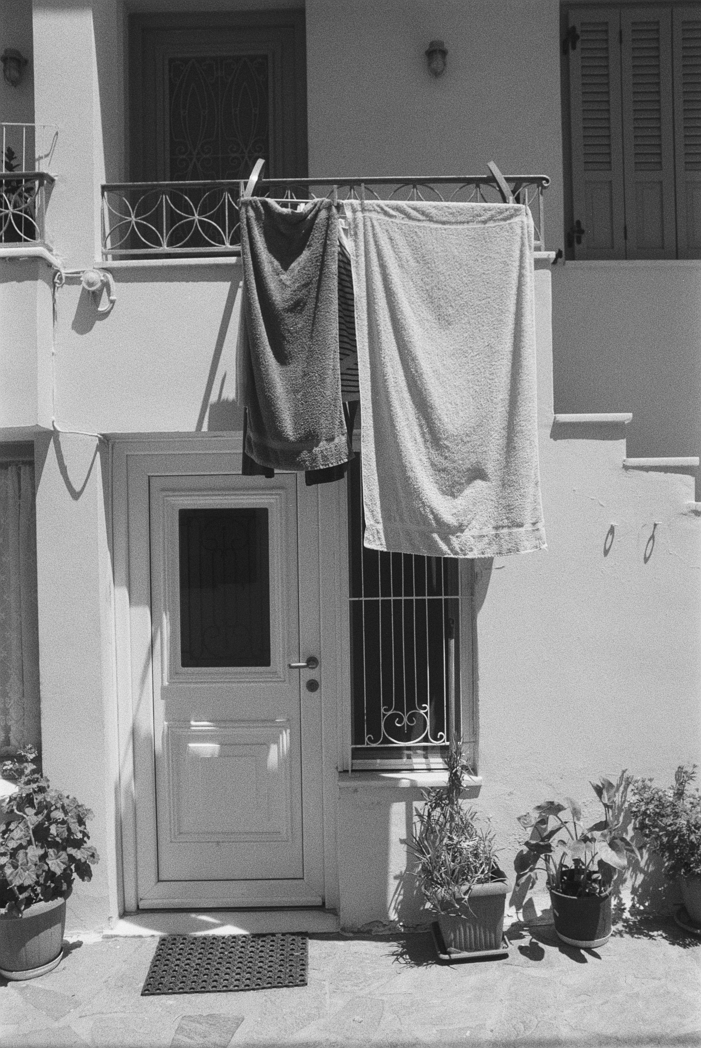 Ein Eingang mit Tür, Fensterläden und einem kleinen Balkon. Es hängen Handtücher zum Trocknen auf einer Wäscheleine. Es gibt mehrere Pflanzen in Töpfen vor dem Eingang.