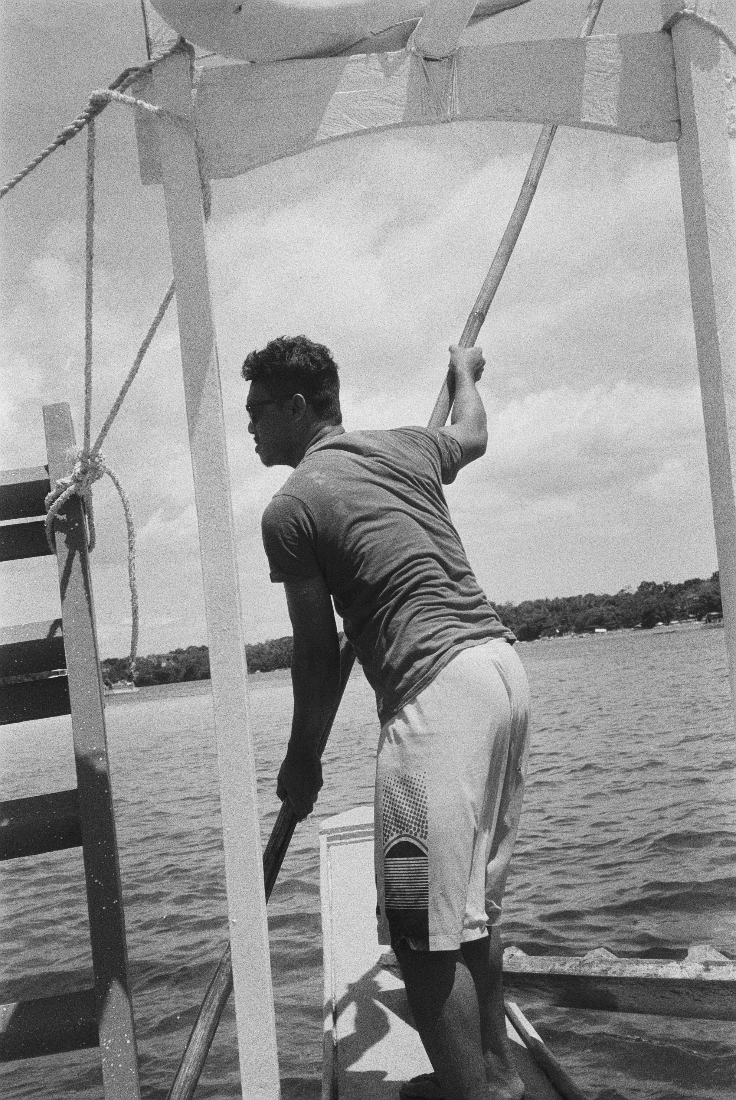Ein junger Mann steht auf einem Boot und hält ein Paddel, während er auf einem Fluss oder See steht. Er trägt eine Kurzarm-Hemd und Shorts. Im Hintergrund sind Wasser, Bäume und ein Himmel mit Wolken zu sehen.