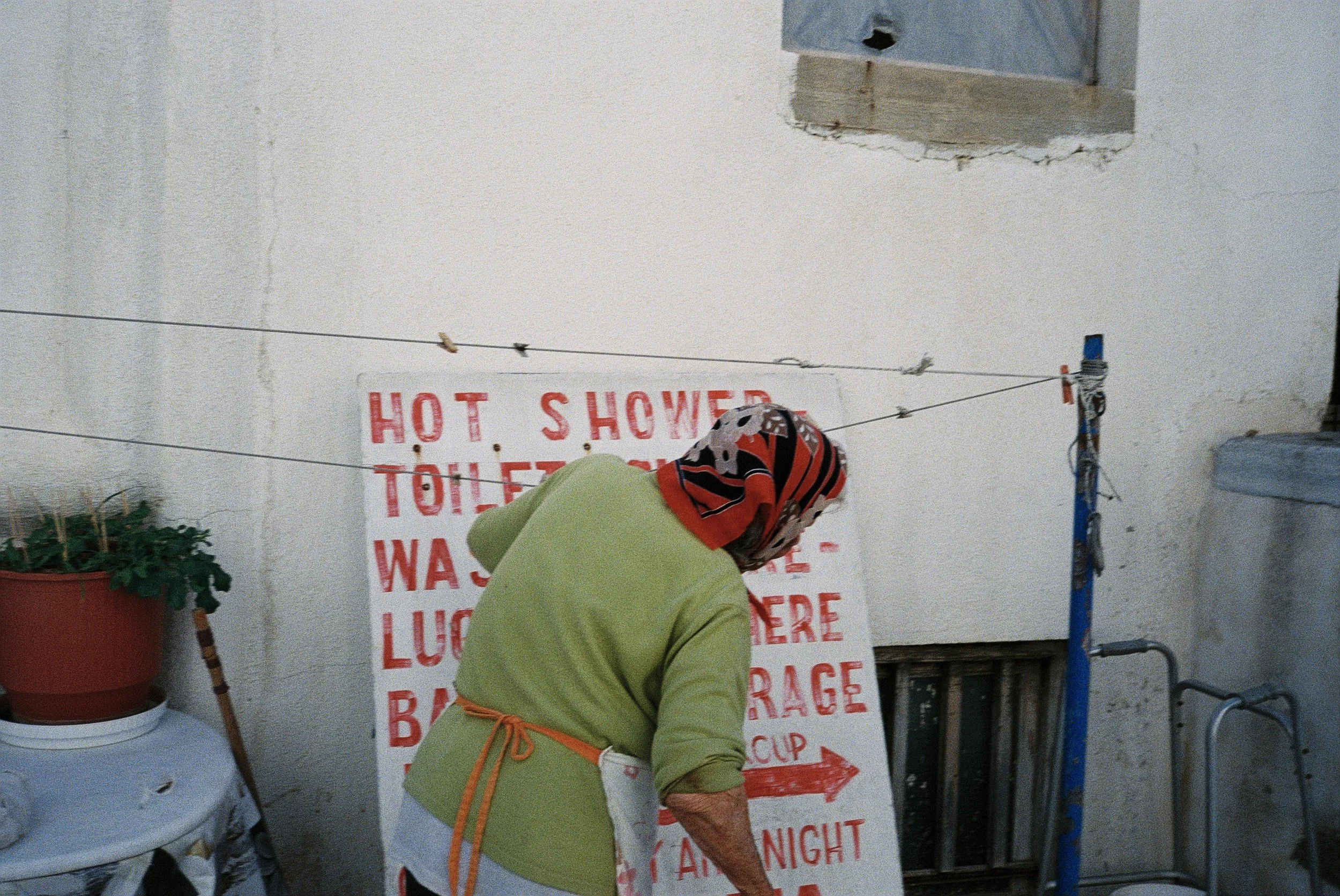 Eine Person trägt einen grünen Mantel und einen roten Schal. Sie stehen vor einem weißen Schild mit roten Buchstaben, das „Hot Shower Toilets Warning” und einen roten Pfeil zeigt. Im Hintergrund ist eine weiße Wand mit einem blauen Wasserhahn und ein