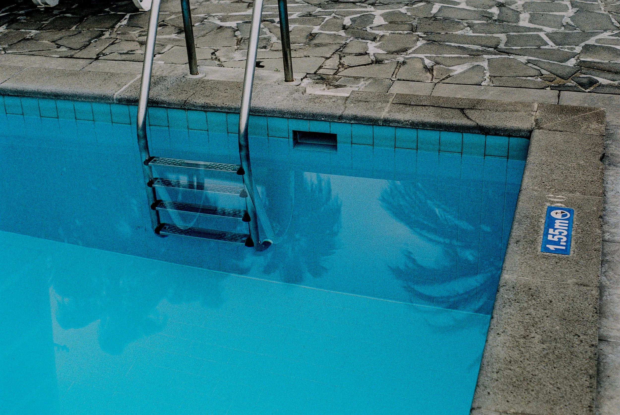 Blick auf einen Swimmingpool mit blauer Wasserfarbe, eine Leiter mit drei Stufen und einem Begrenzungsstreifen mit einer Wasserstandmarkierung bei 1,55 Metern. Der Pool ist von einem gepflasterten Bereich umgeben.
