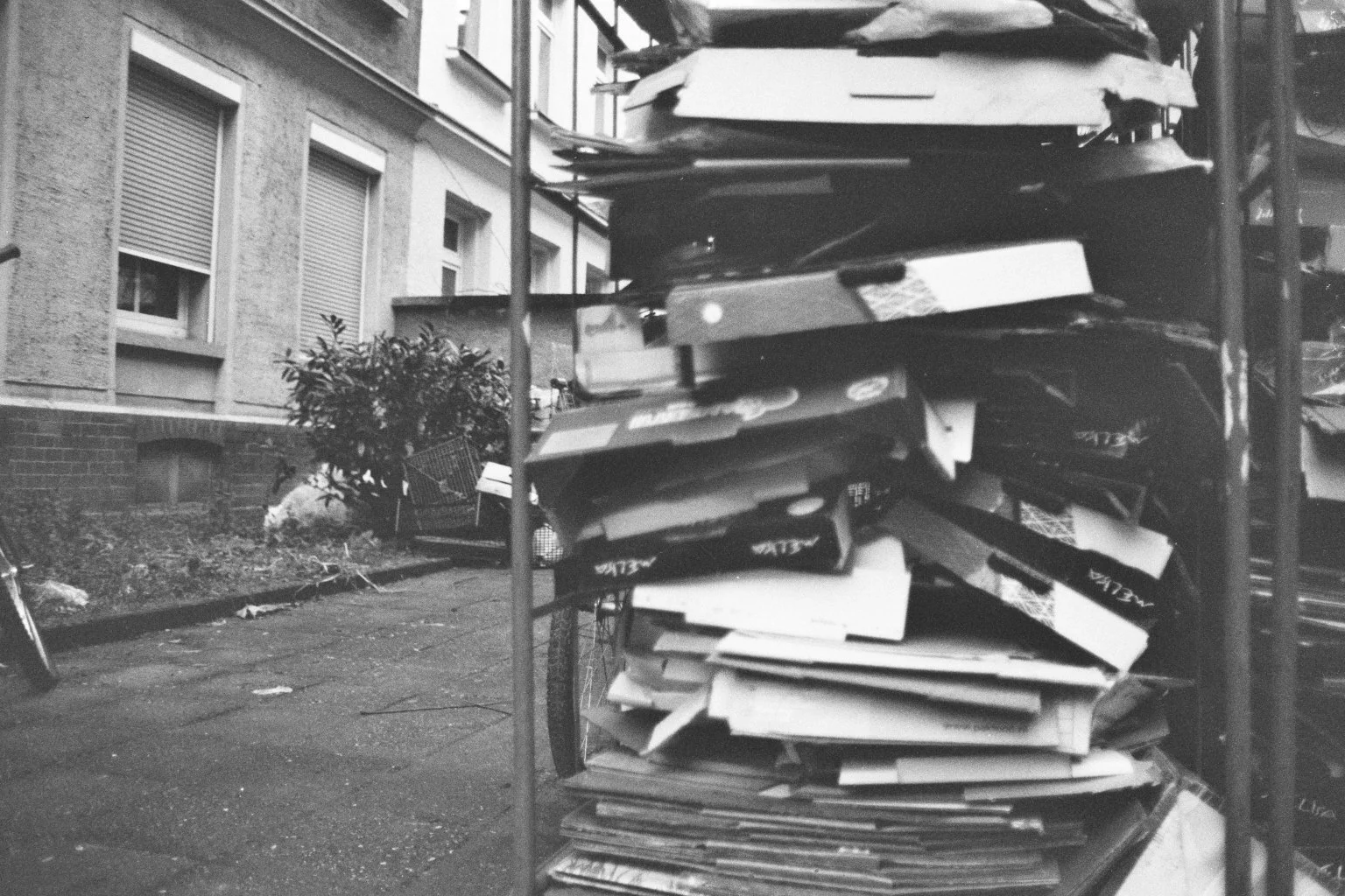 Schwarzer metalischer Bücherwagen mit gestapelten Büchern und Papieren, im Hintergrund eine Hausfassade mit Fenstern und einer kleinen Hecke