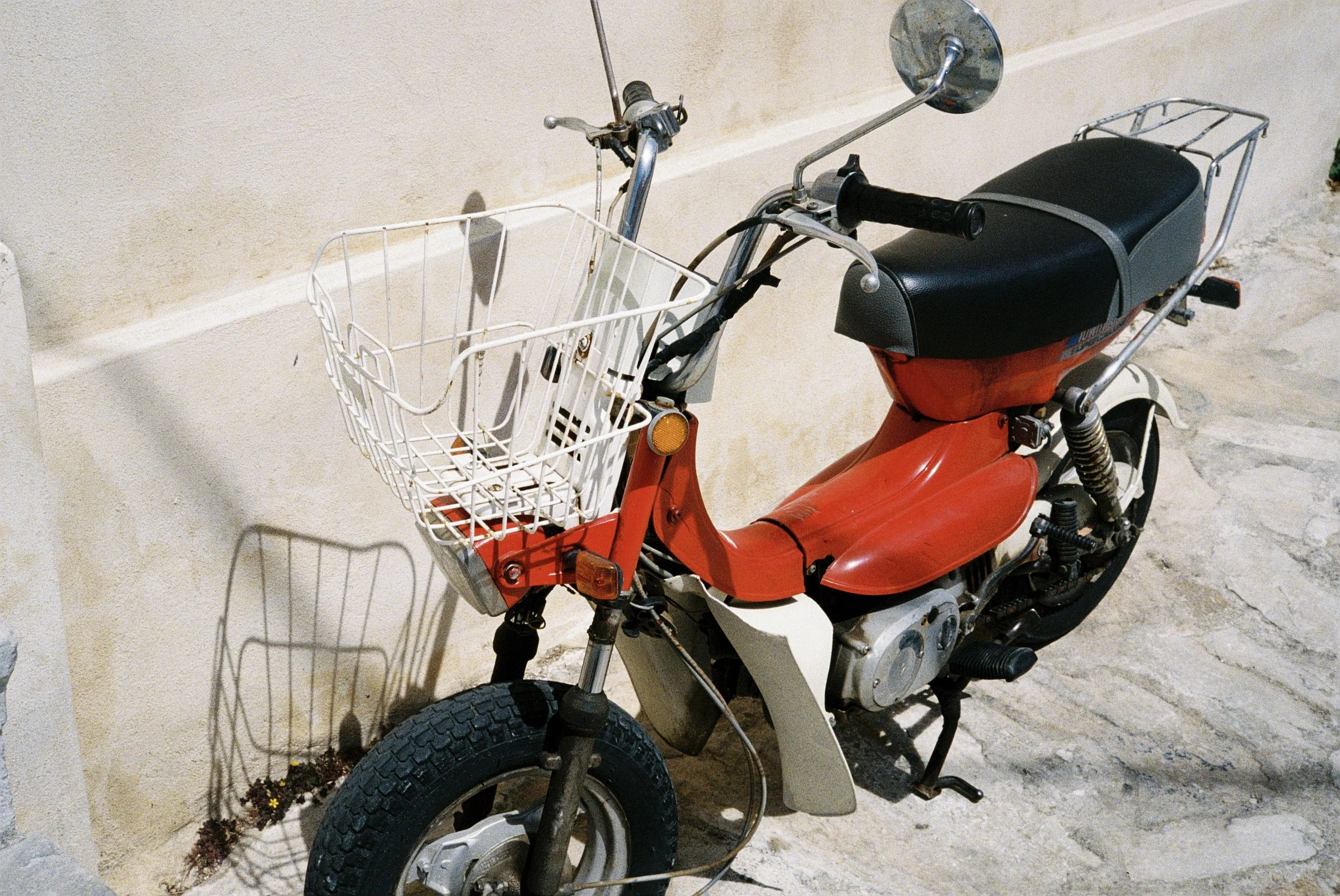 Ein rotes Moped mit einem Einkaufswagenkorb vorne, das gegen eine weiße Wand steht.