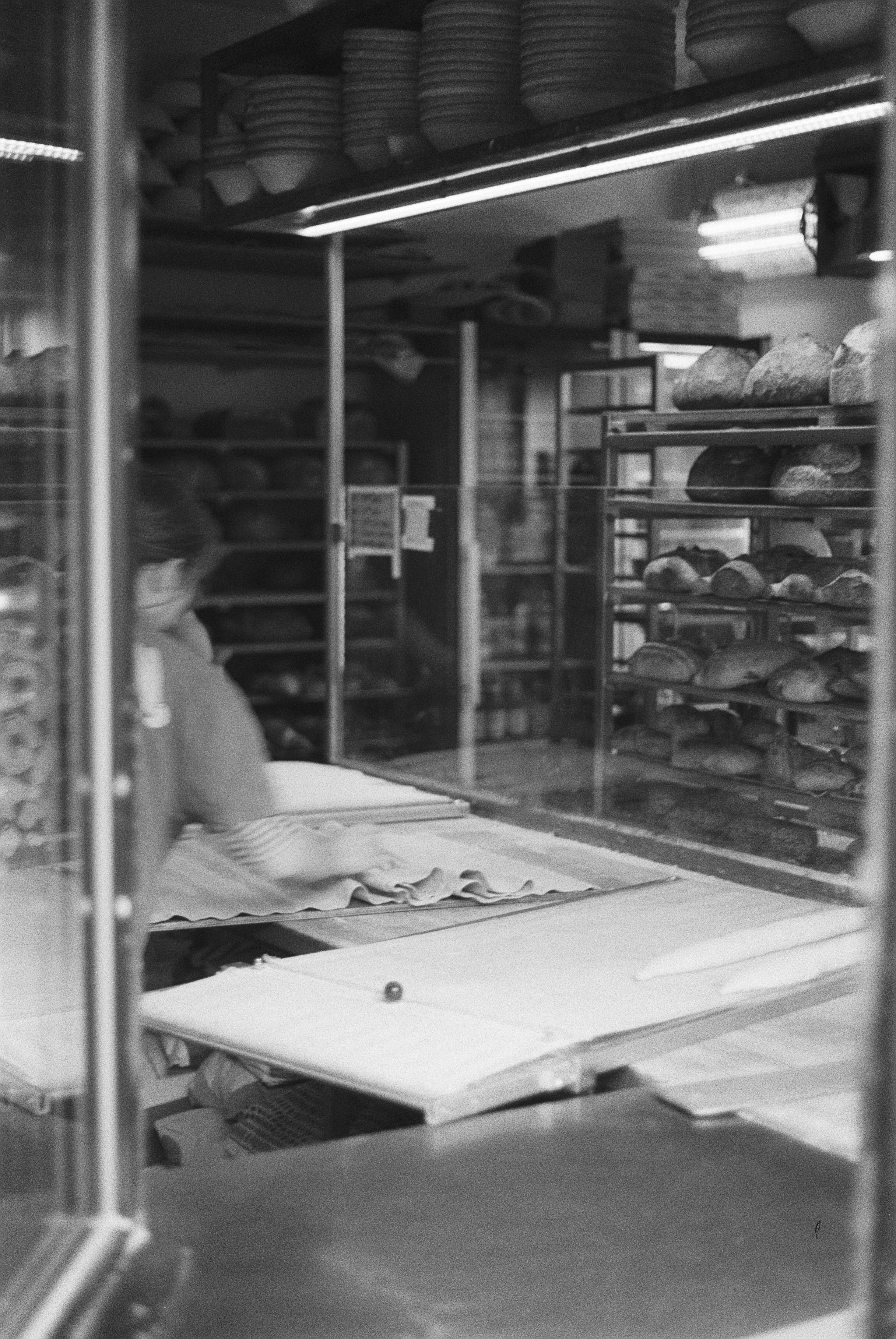 Blick durch eine Glasscheibe in eine Bäckerei, in der Brote auf Regalen in der Backstube gelagert werden.