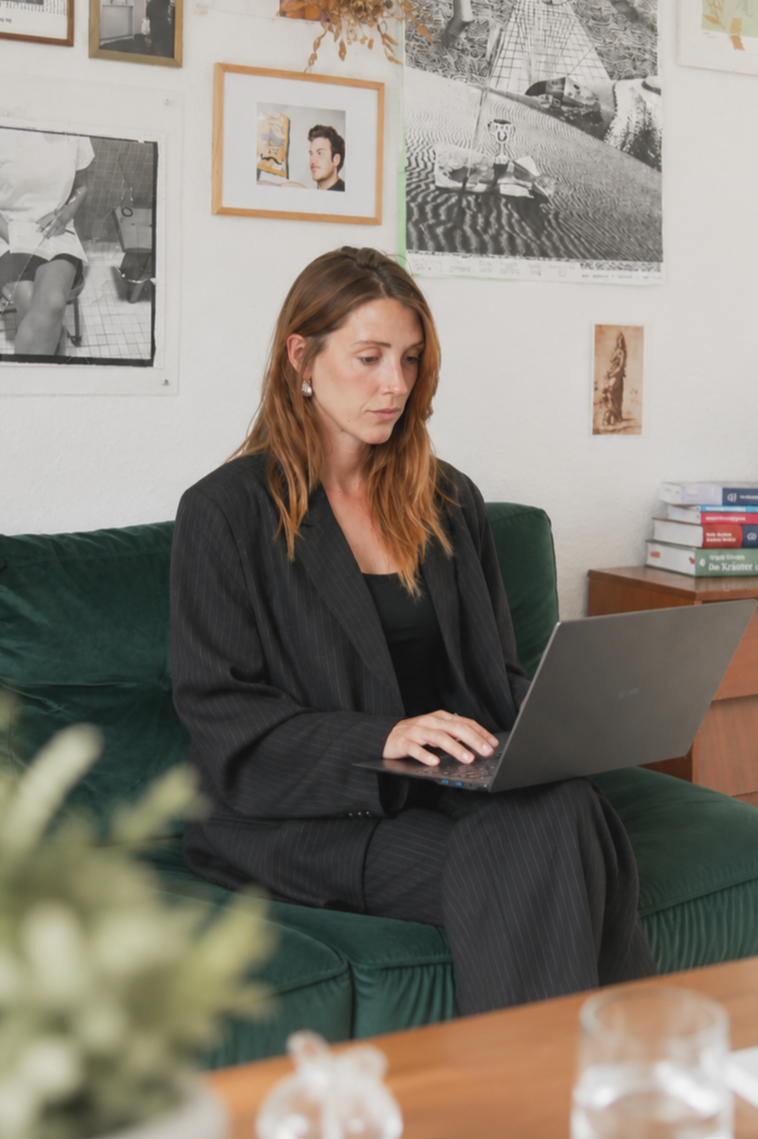 Frau in schwarzem Anzug arbeitet an einem Laptop auf einer grünen Couch in einem Raum mit Kunstwerken an der Wand.