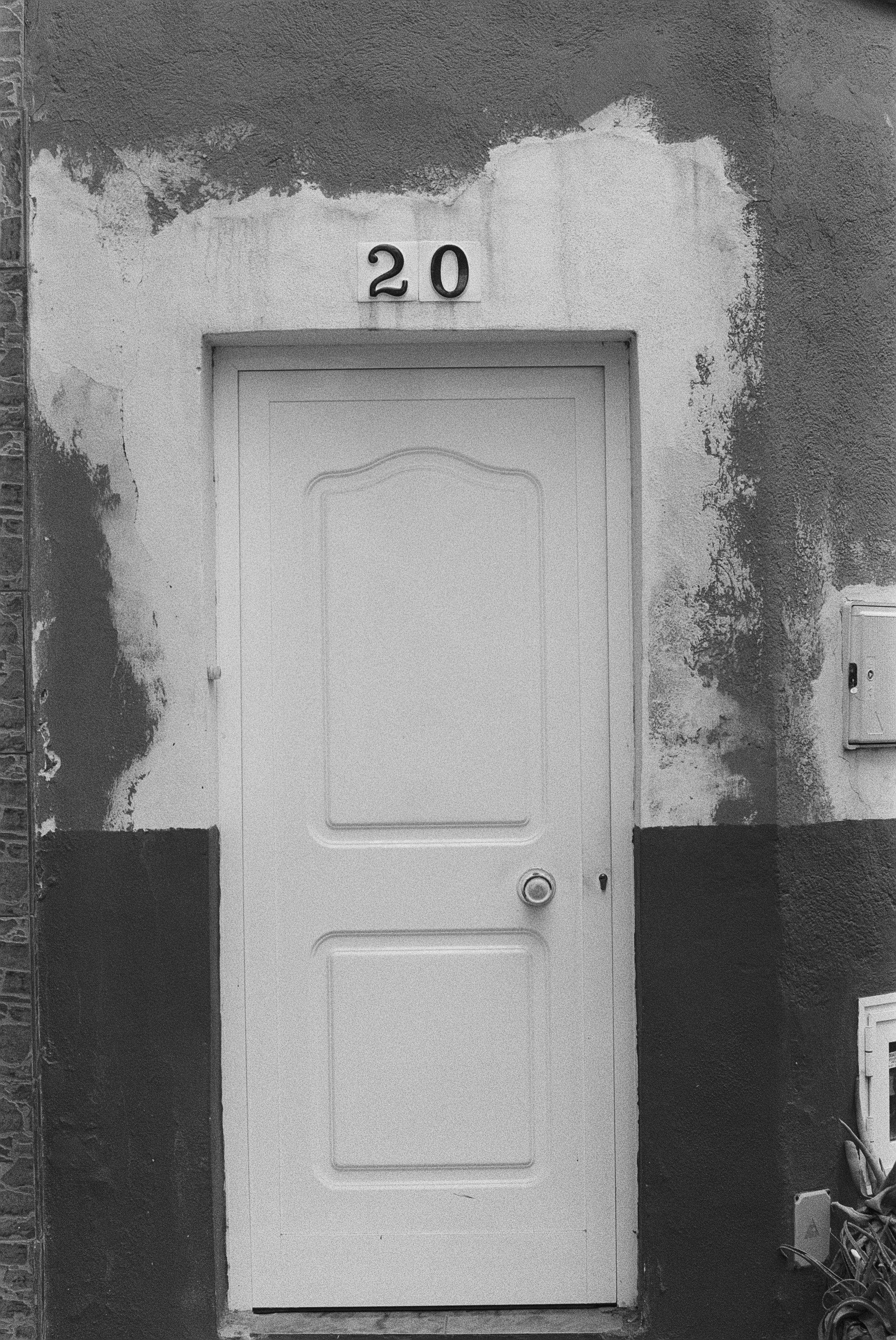 Weiße Tür mit der Hausnummer 20 an einer verputzten Wand mit abblätternder Farbe, im Eingangsbereich, im Schwarz-Weiß-Foto.
