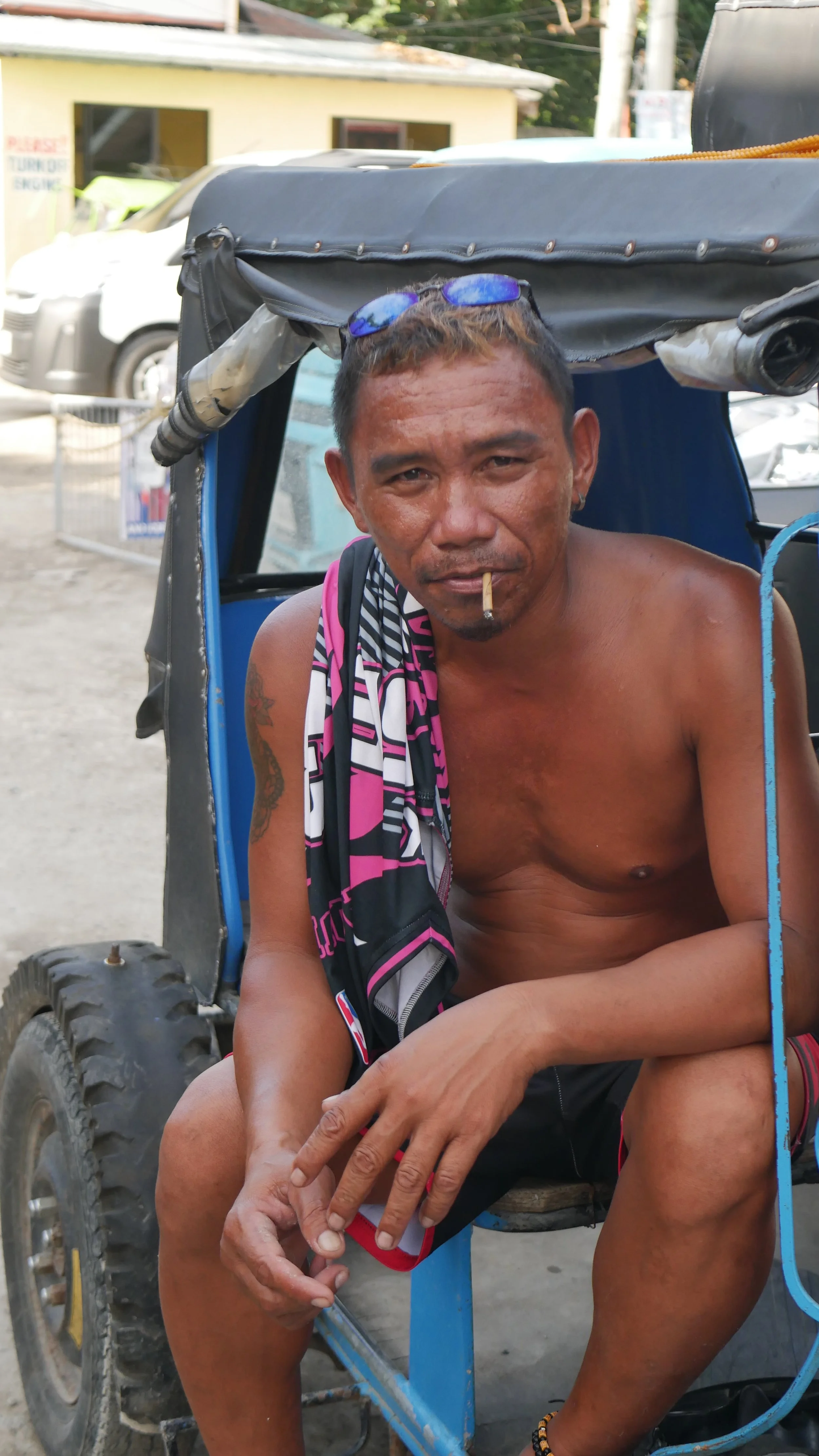 Ein Mann sitzt in einem blauen Tuk-Tuk, ohne Hemd, raucht eine Zigarette und trägt eine schwarze und pinkfarbene Sportjacke über der Schulter.
