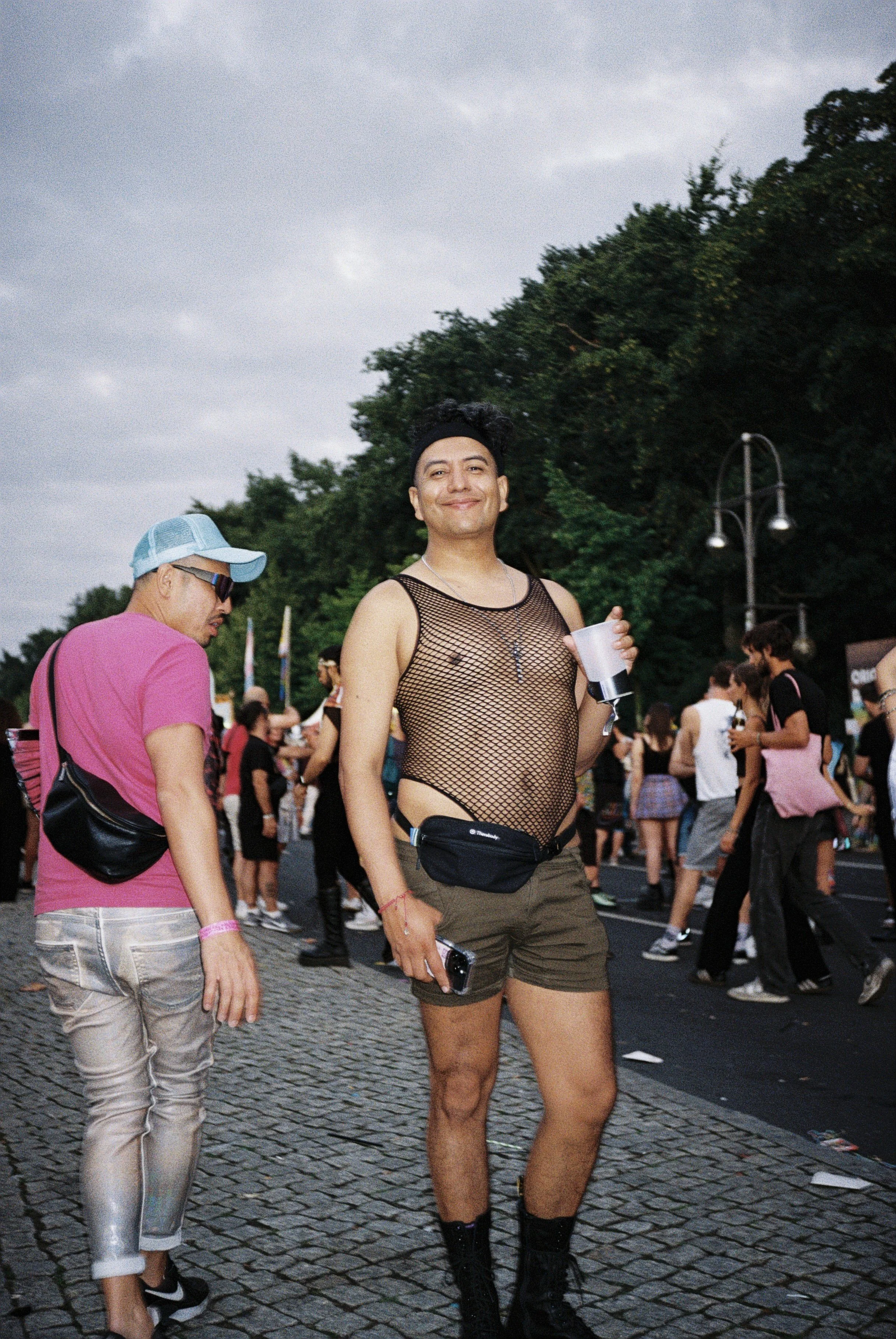 Ein junger Mann trägt eine schwarze Netzhemden, braune Shorts, schwarze Stiefel und eine schwarze Gürteltasche, während er in einem Menschenmengen bei einer Veranstaltung im Freien steht.